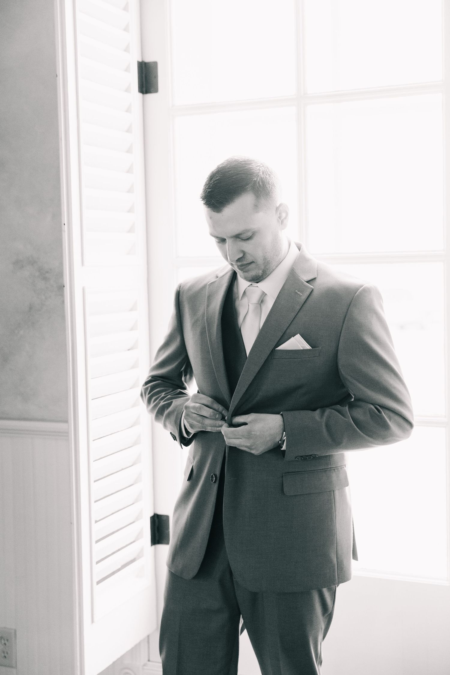 Groom in gray suit adjusts jacket button in black and white portrait by window with shutters.