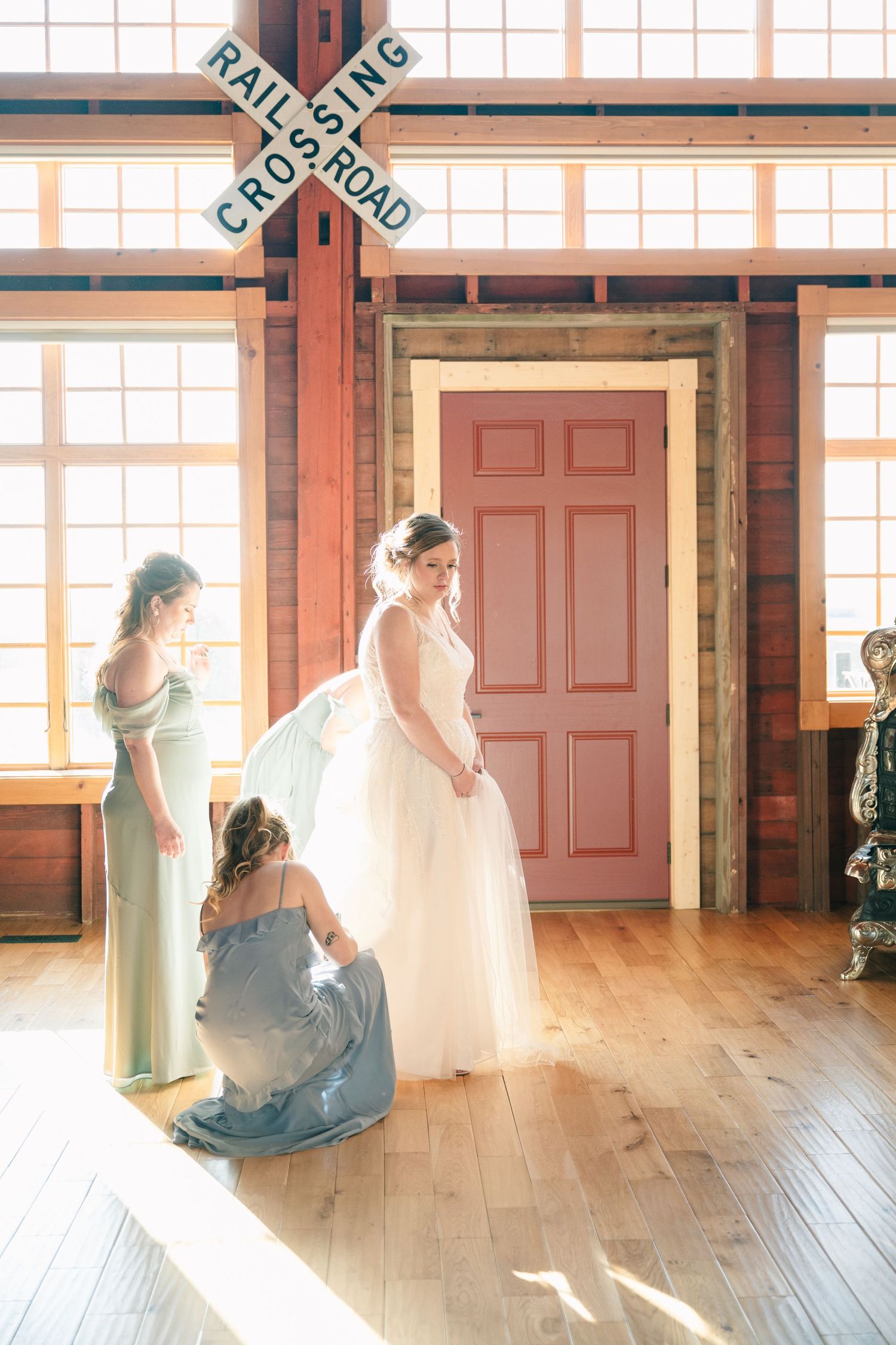 Bridesmaids helping bride with her gown inside a sunlit rustic venue with a railroad crossing sign on the wall.