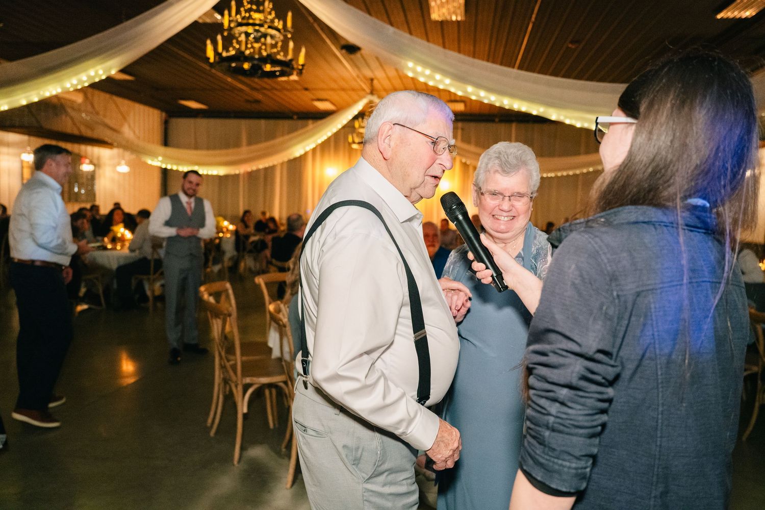 Elderly couple dances joyfully while being interviewed with microphone at warmly lit wedding reception.