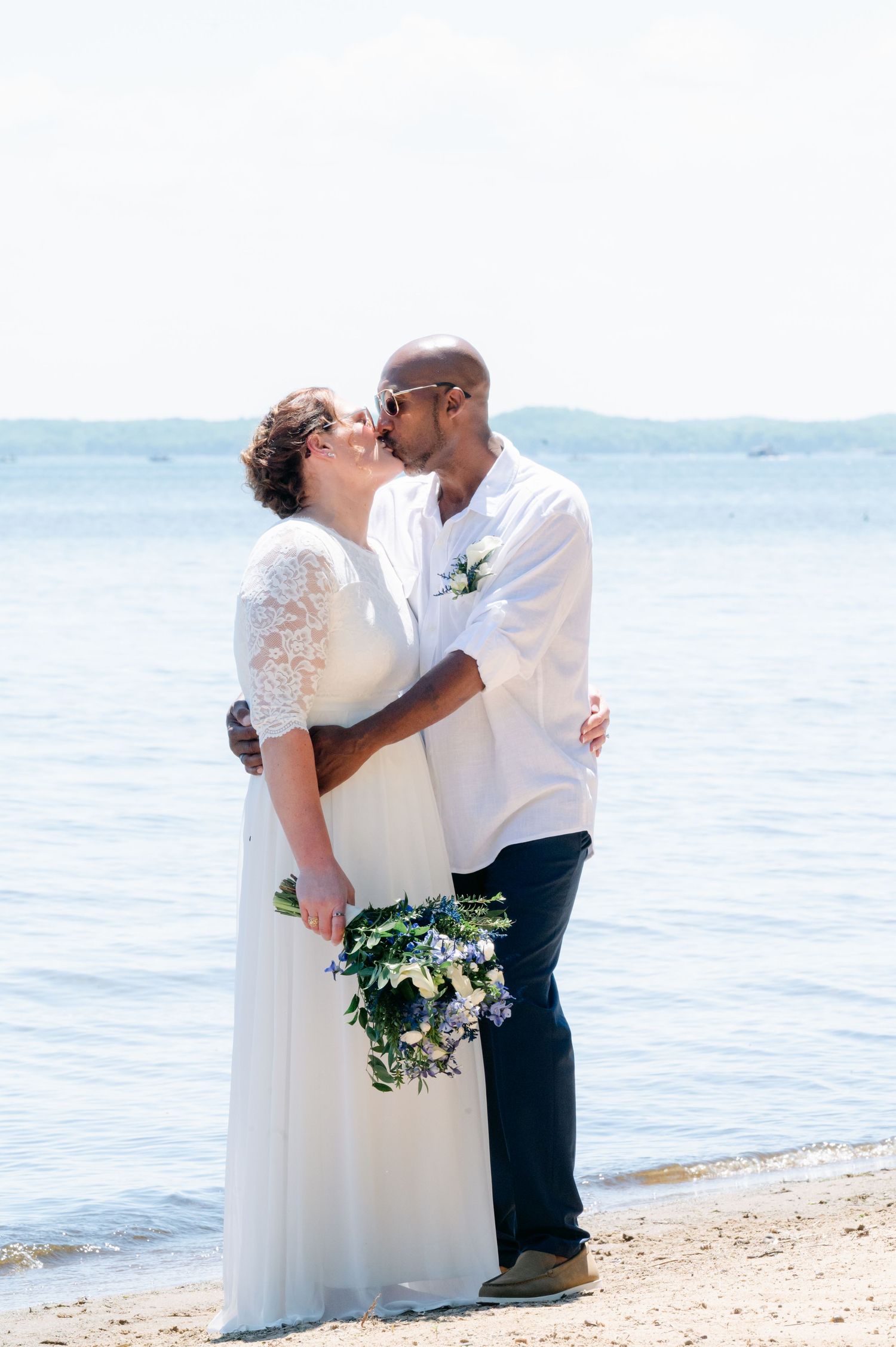 Bride and groom sharing a romantic kiss on a sandy beach with calm blue water behind them.