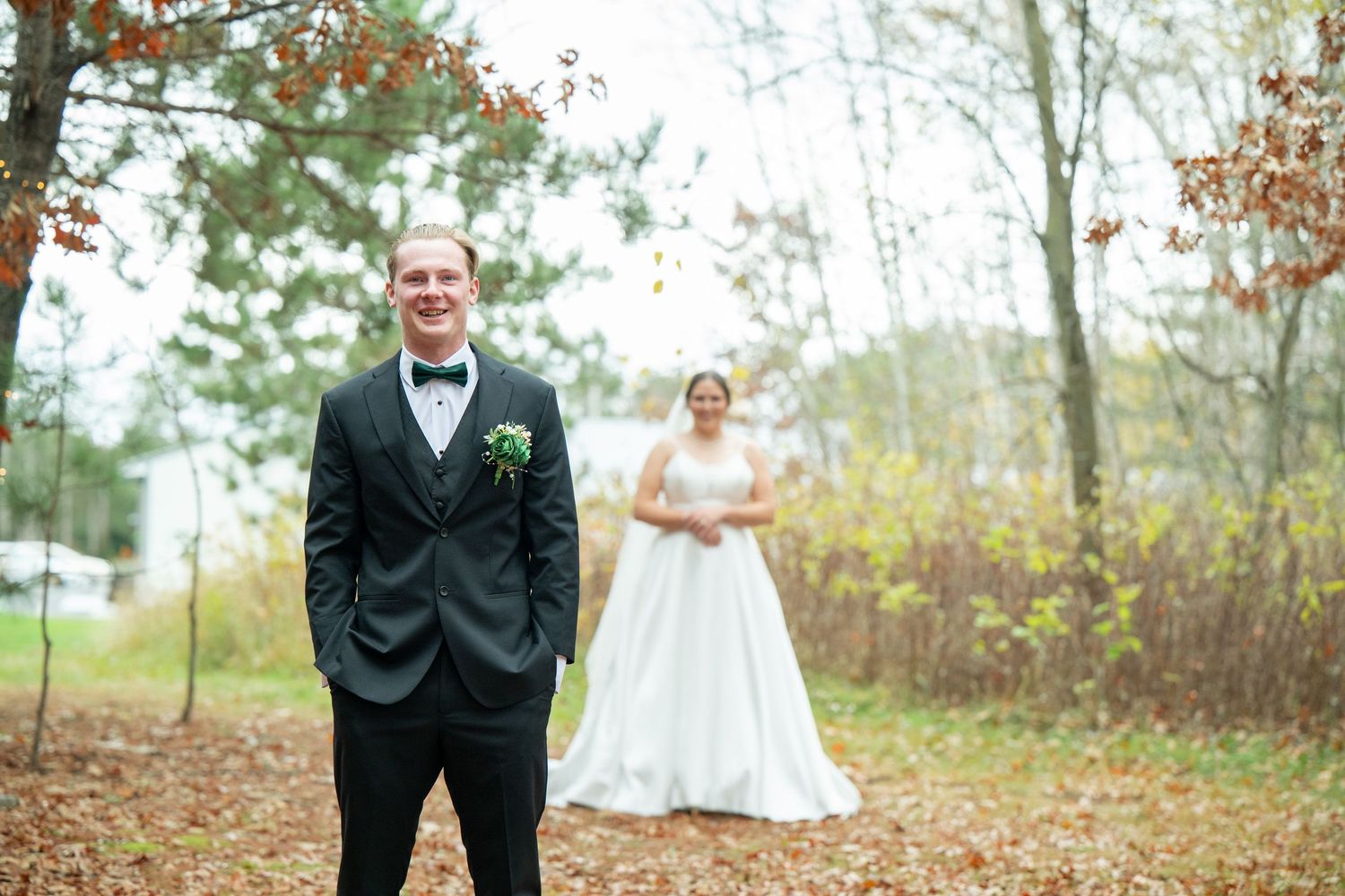 Newlyweds stand together in a wooded autumn setting with fallen leaves scattered on the ground.
