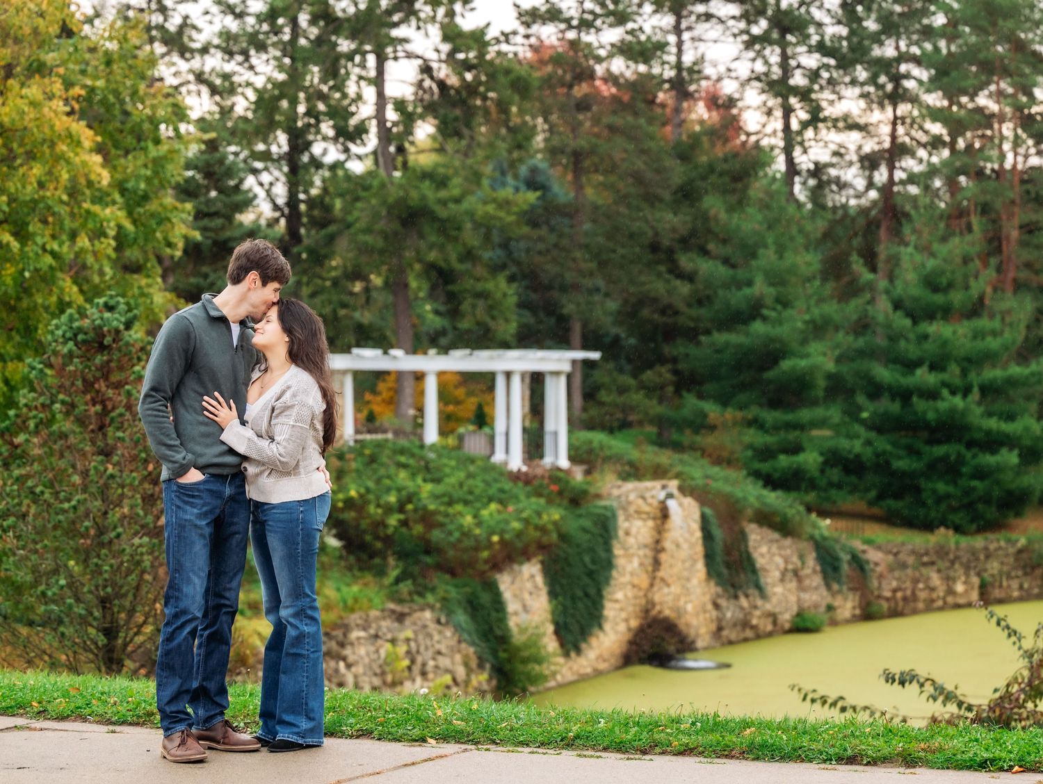 A couple stands together on a stone wall pathway surrounded by autumn trees and landscaping at Lake Como. Engagement Sessions Minnesota.