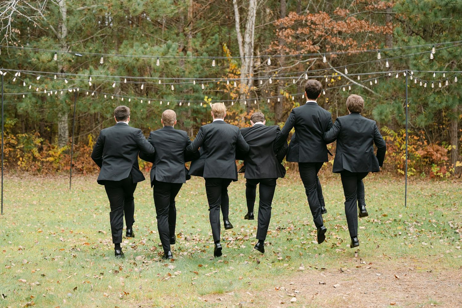 Groomsmen in black suits pose playfully from behind in outdoor wedding photo.