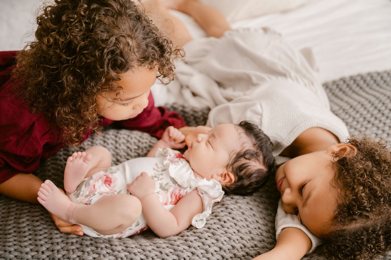 Three people with curly hair lie together on a gray blanket, sharing a tender family moment.