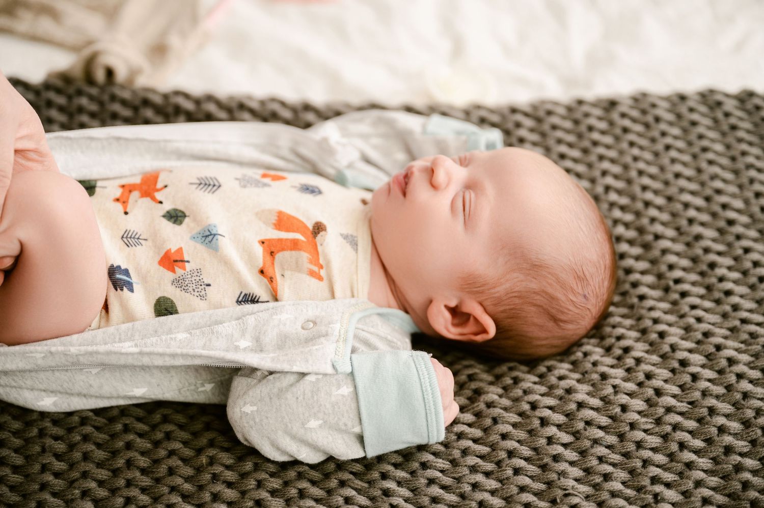 Tiny baby wearing a white onesie with fox print design rests peacefully on gray knitted blanket.