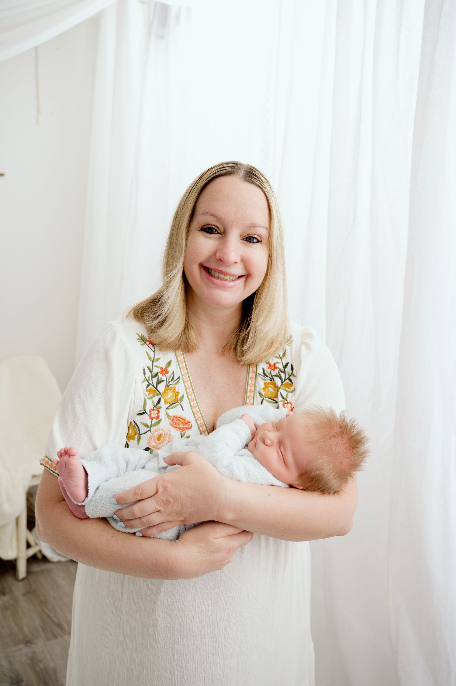 A tender moment captured as a parent in a white embroidered dress holds their sleeping newborn baby in a bright room.