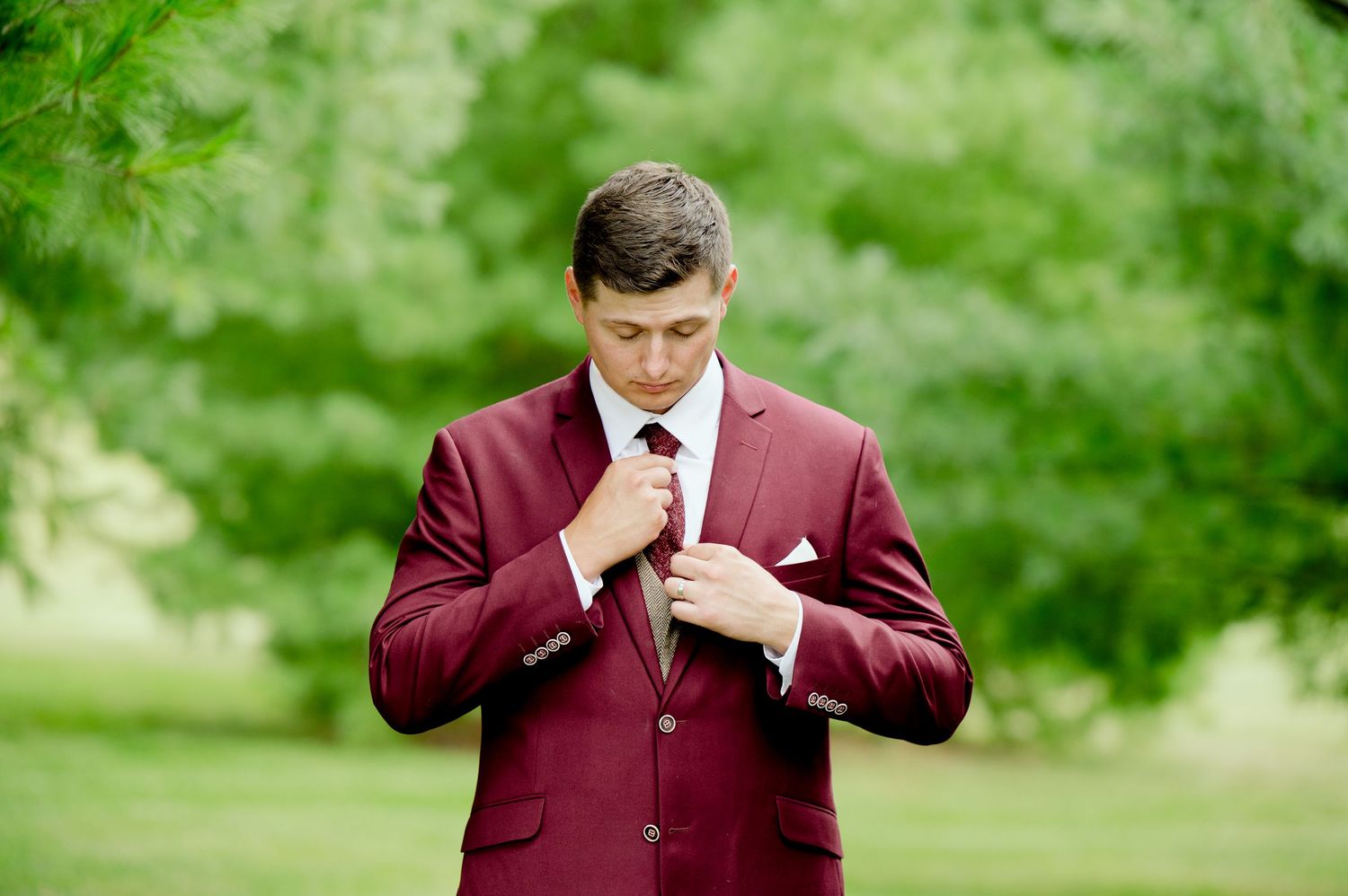 A groom in a burgundy suit adjusts his tie in a series of outdoor wedding portraits.