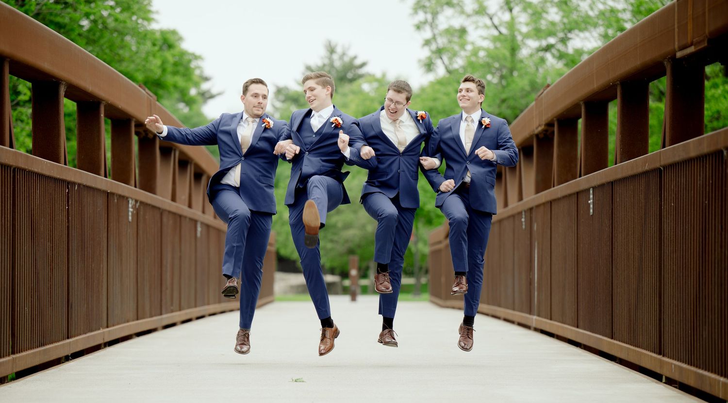 Four groomsmen in navy suits jumping in synchronized pose on wooden bridge surrounded by greenery.