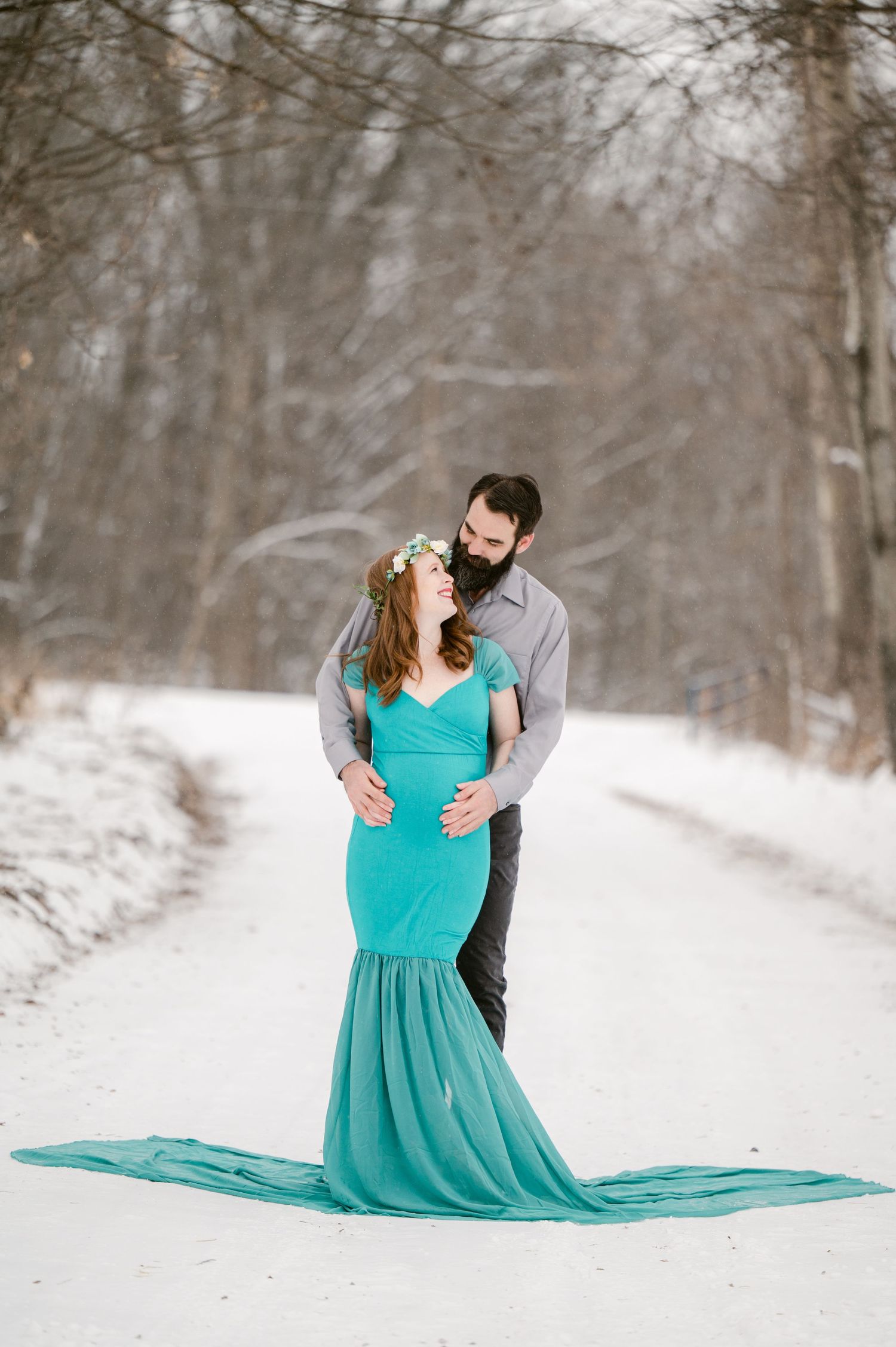 Winter couple photoshoot with figure in flowing turquoise dress against snowy forest backdrop.
