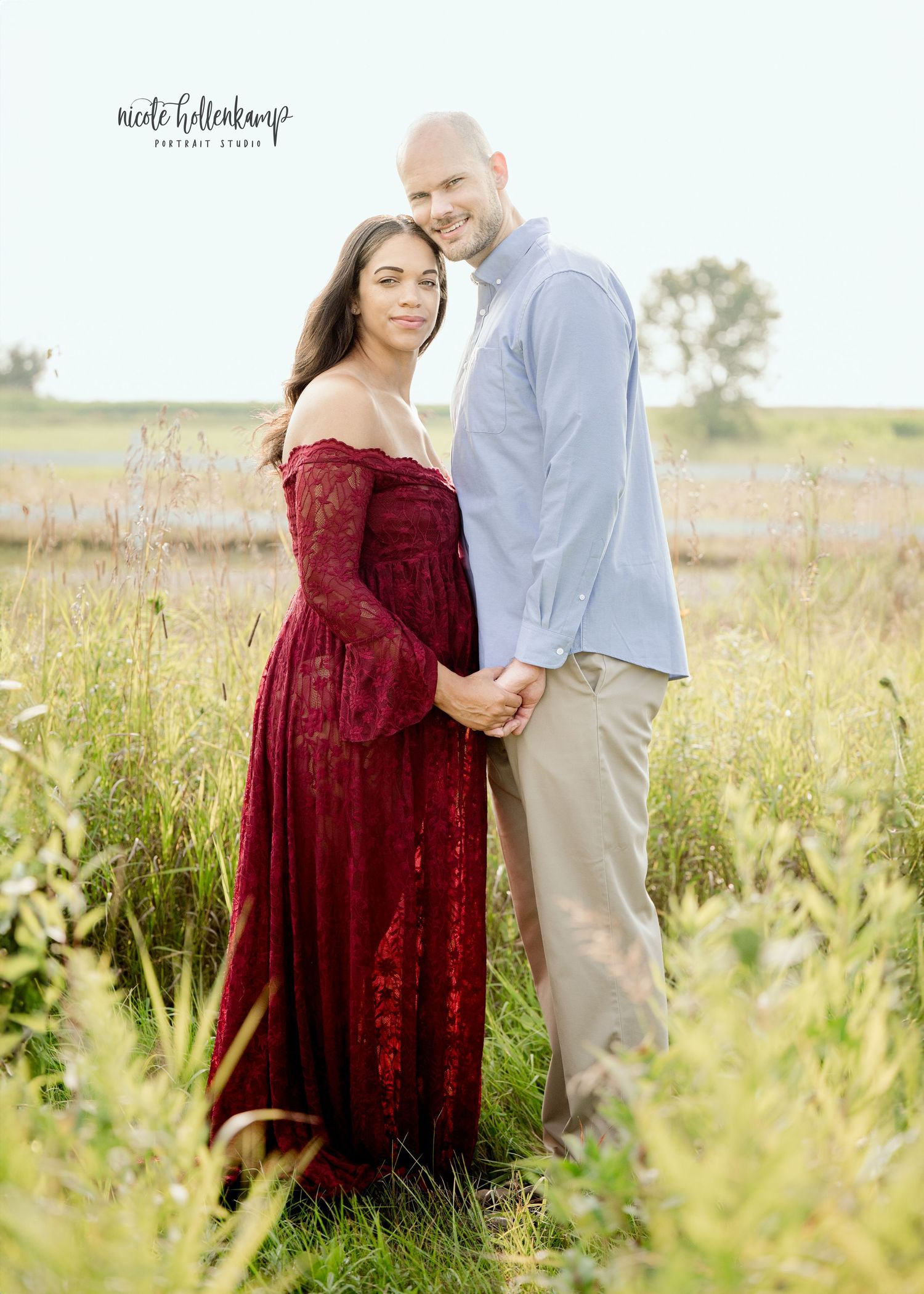 Pregnant couple posing in a field, woman in red lace gown, man in blue shirt, maternity portrait session.