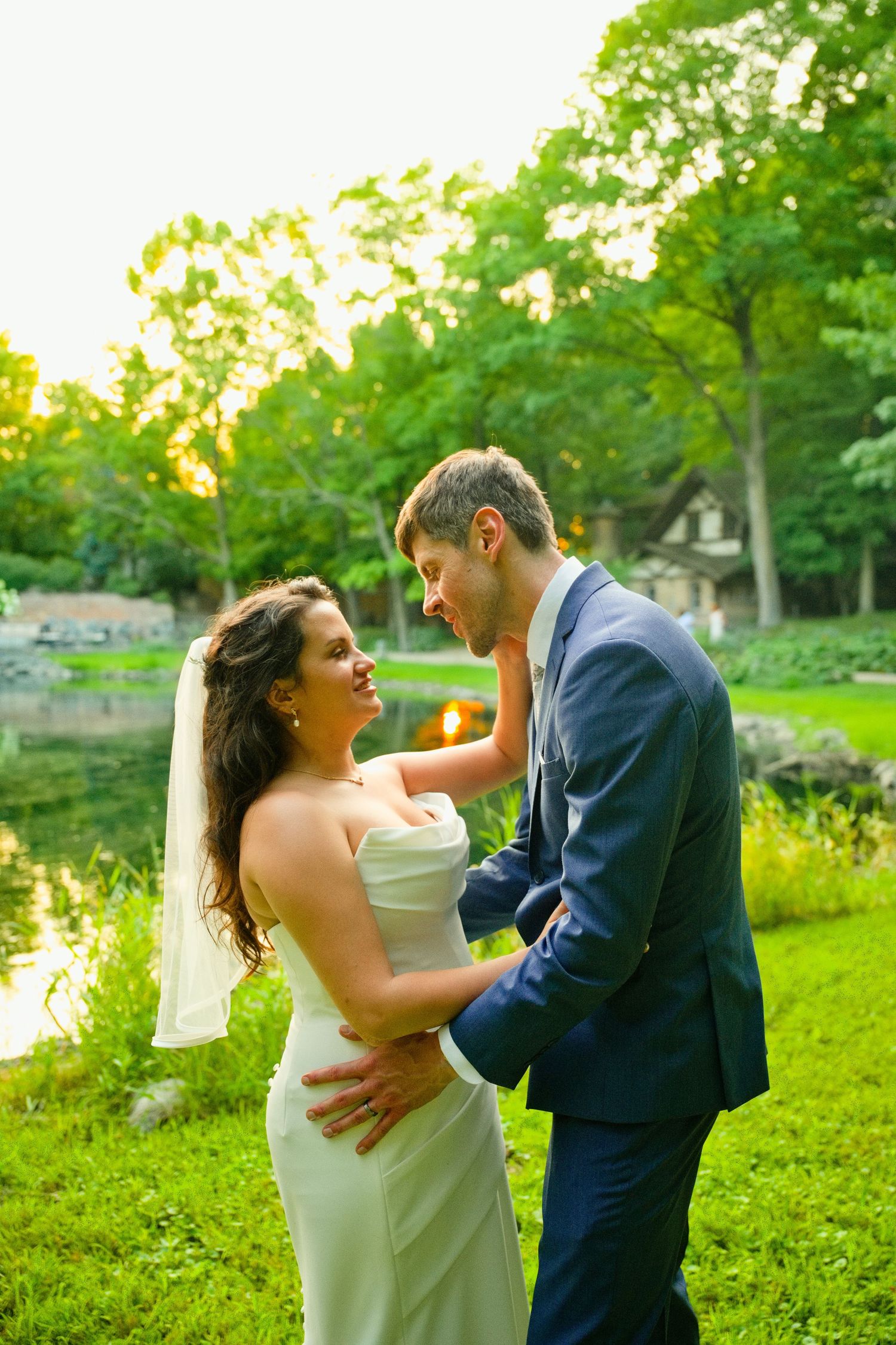 Bride and groom share a romantic moment by a pond in a lush green park at sunset.