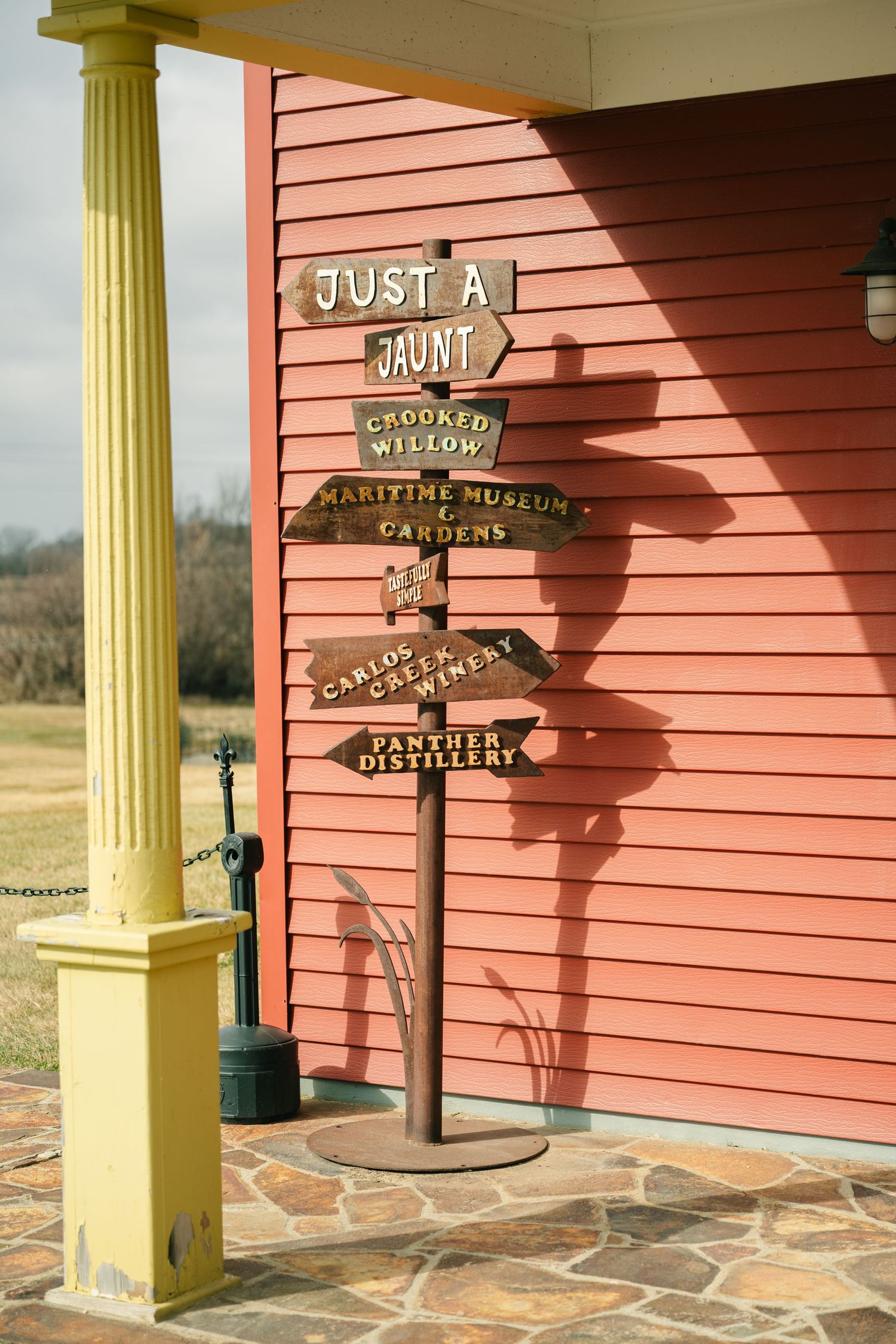 Rustic wooden directional sign reading Just A Jaunt with local attraction arrows beside a red building and yellow column.