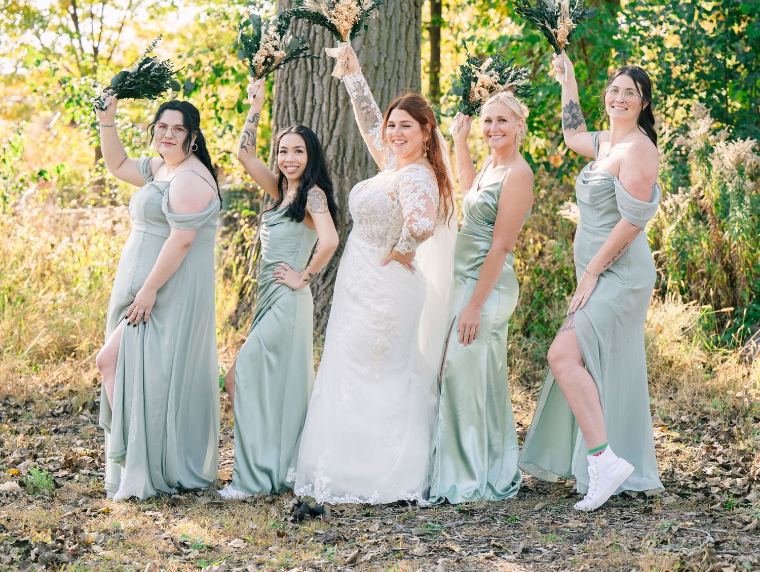 Bride in white lace gown poses with four bridesmaids in sage green dresses outdoors among autumn trees.