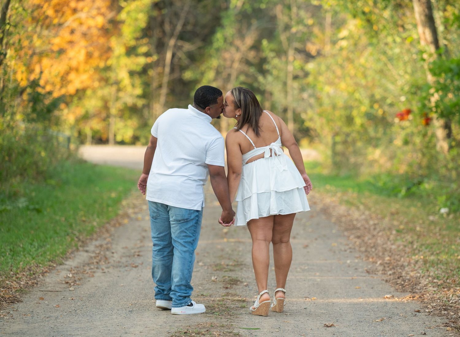 A couple in casual attire share a romantic moment on a dirt path surrounded by autumn foliage.