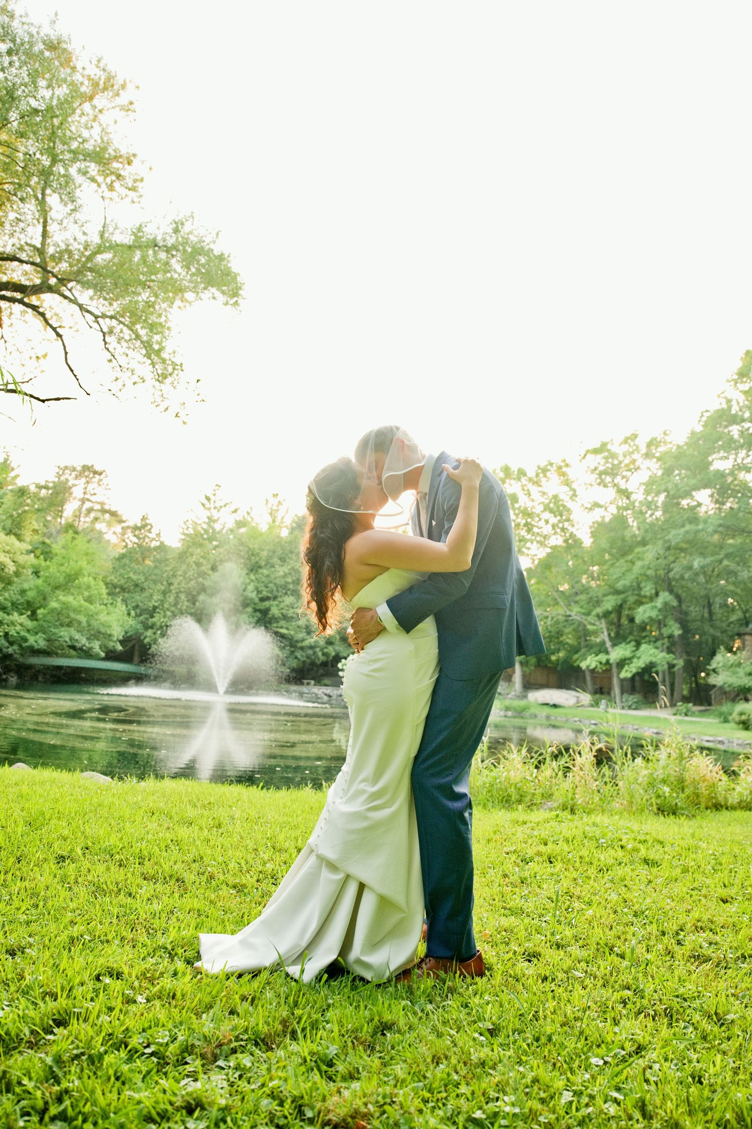 Bride and groom share a romantic kiss on lush green grass near a pond with fountain, surrounded by trees.