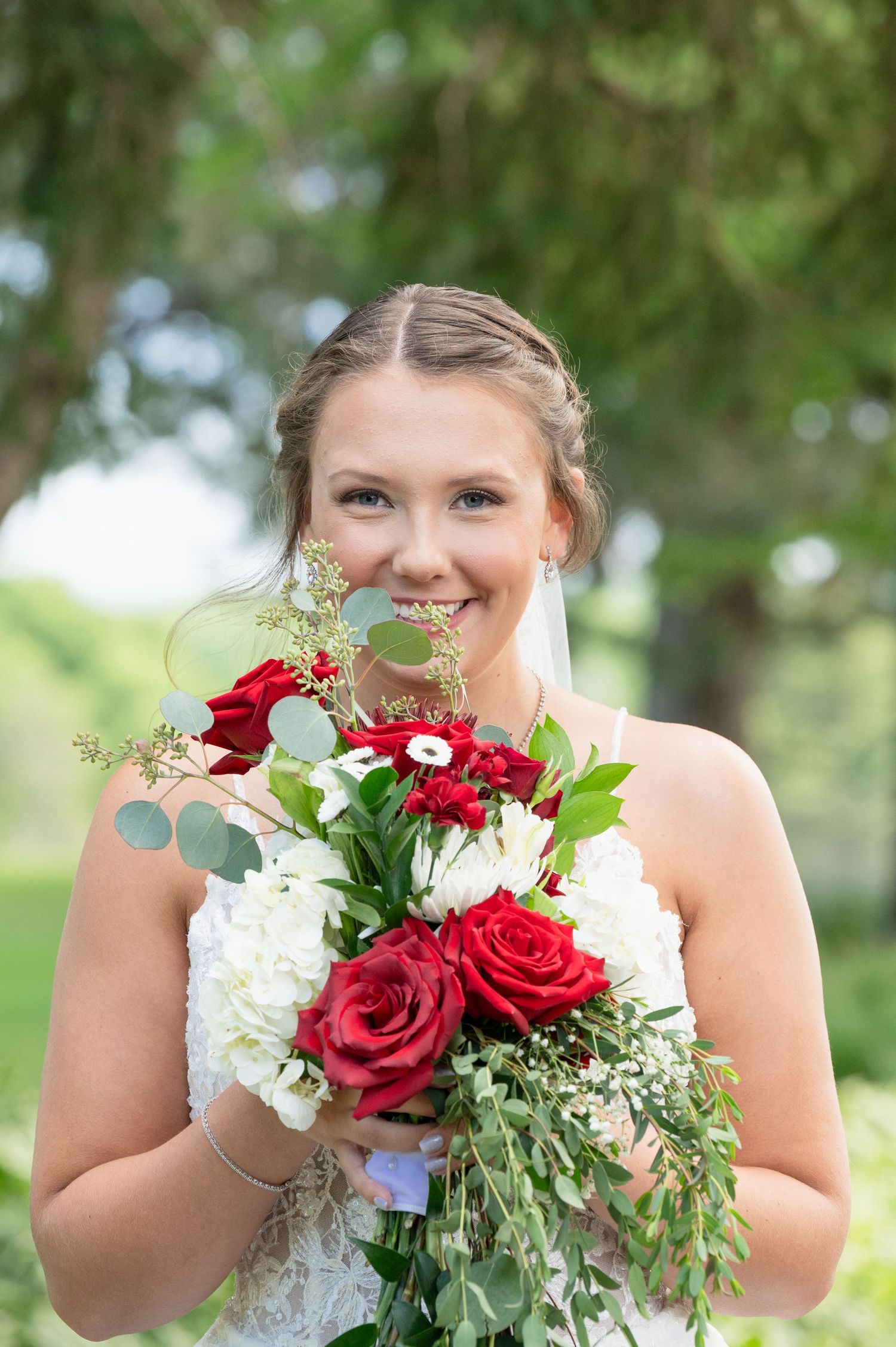 Smiling bride holds a bouquet of red roses and white hydrangeas on her wedding day outdoors.