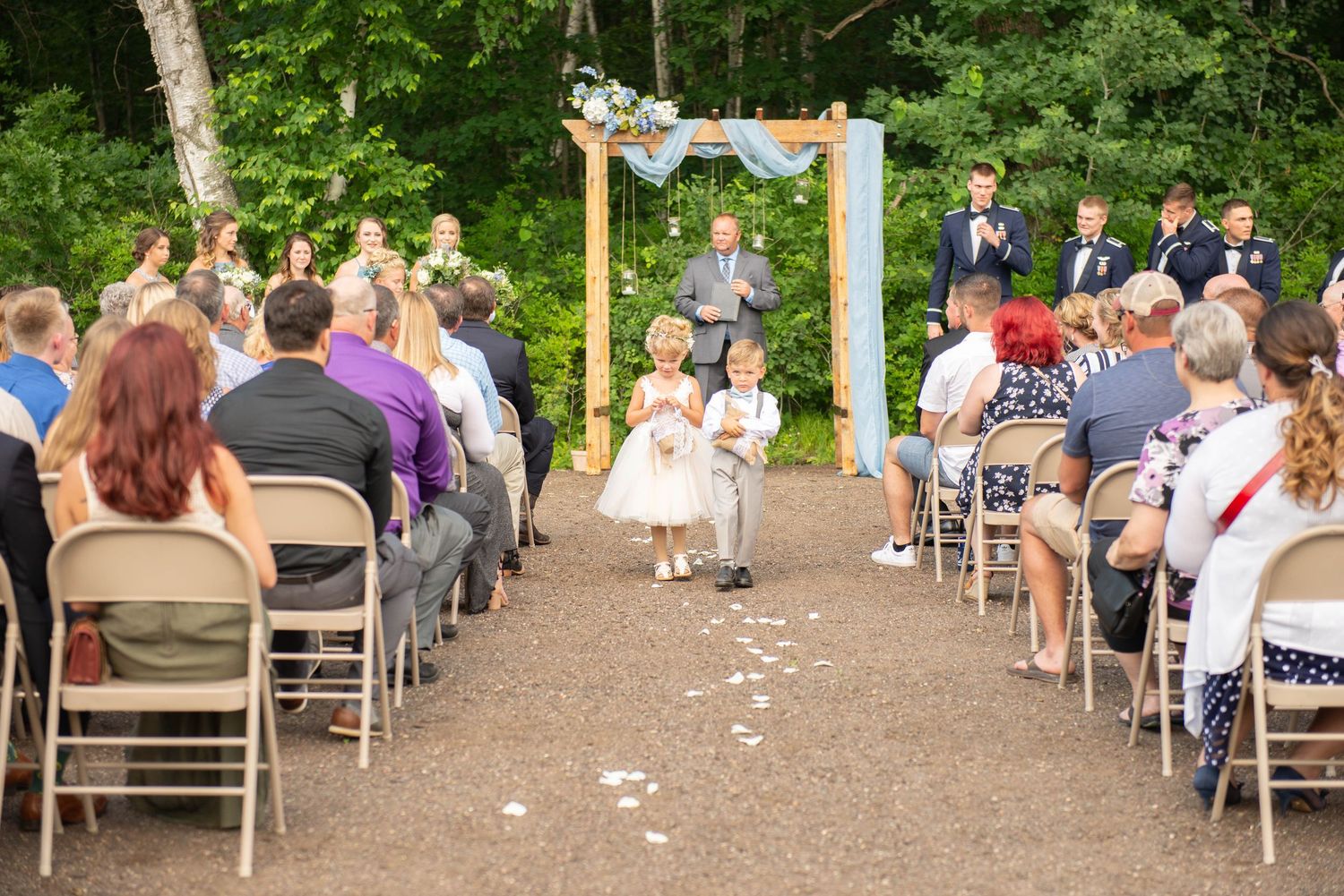Wide view of an outdoor wedding ceremony showing guests seated on wooden chairs with white flower petals scattered on the ground. Zimmerman