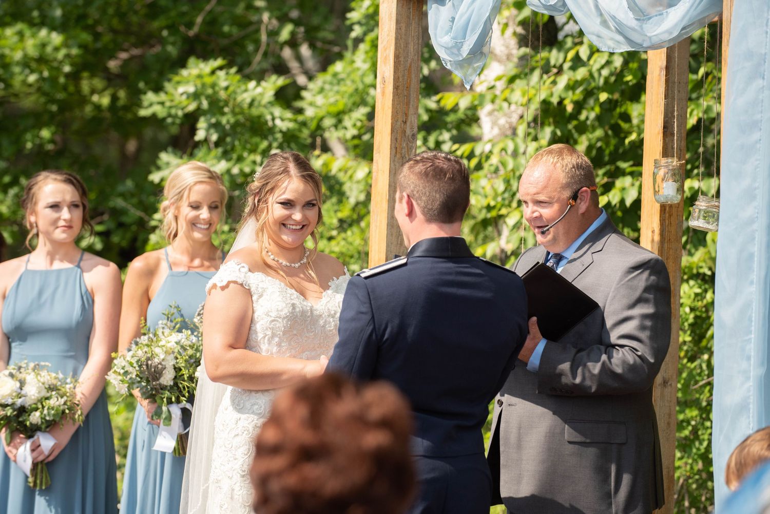Outdoor wedding ceremony with wooden pillars and white fabric draped across, surrounded by green foliage. Zimmerman MN Weddings