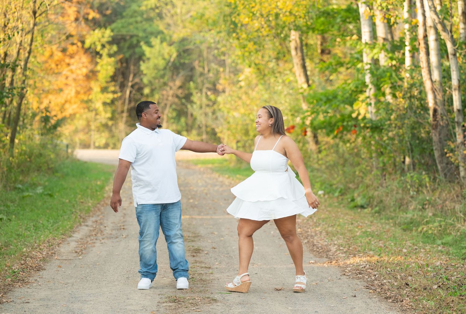 Central Minnesota Best Engagement Photo Couple in white outfits holding hands on a scenic forest path surrounded by colorful autumn foliage.