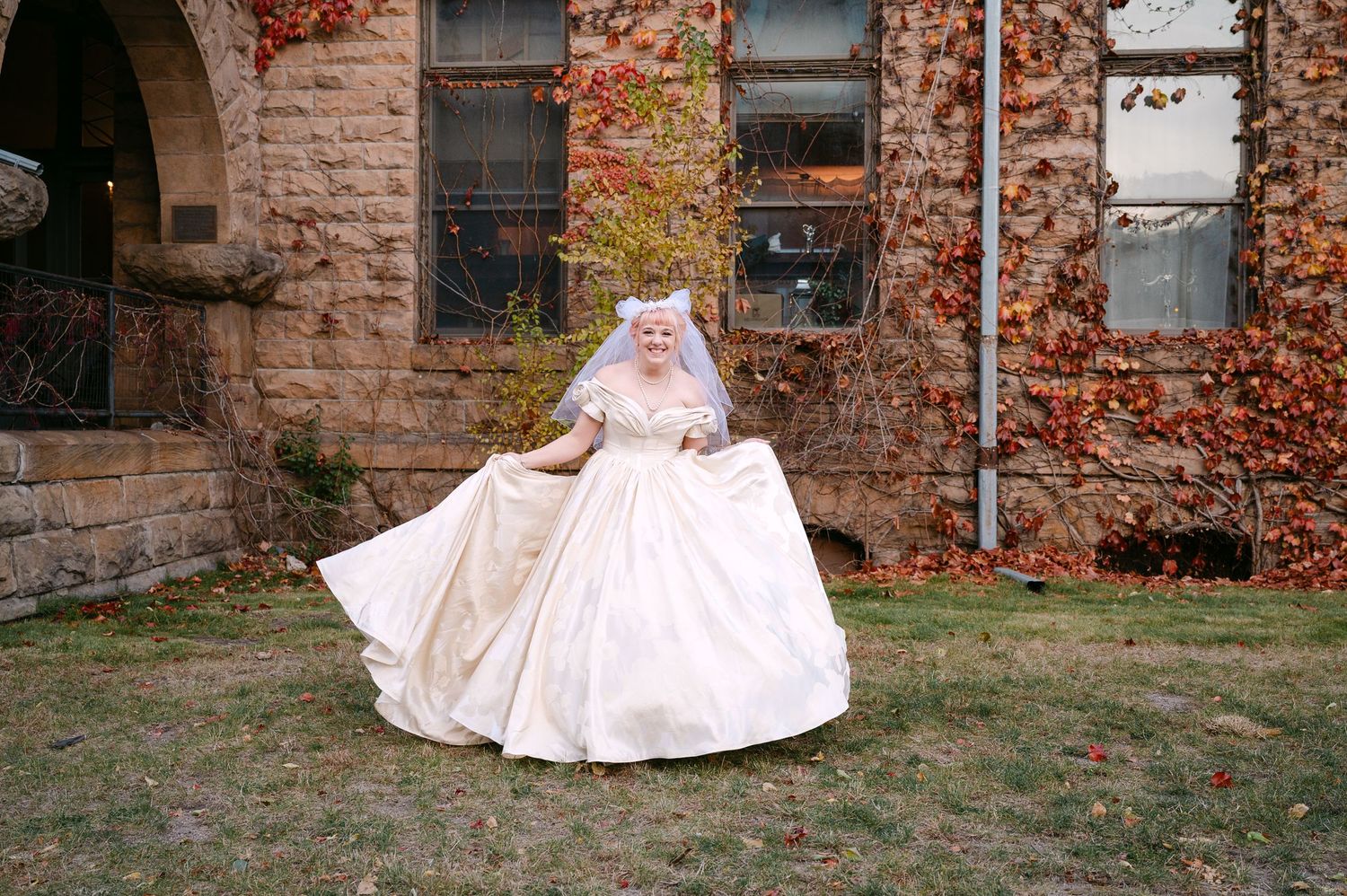 Central MN Wedding Photos | Bride in ivory off-shoulder ballgown and veil twirls outside stone building with autumn ivy backdrop.