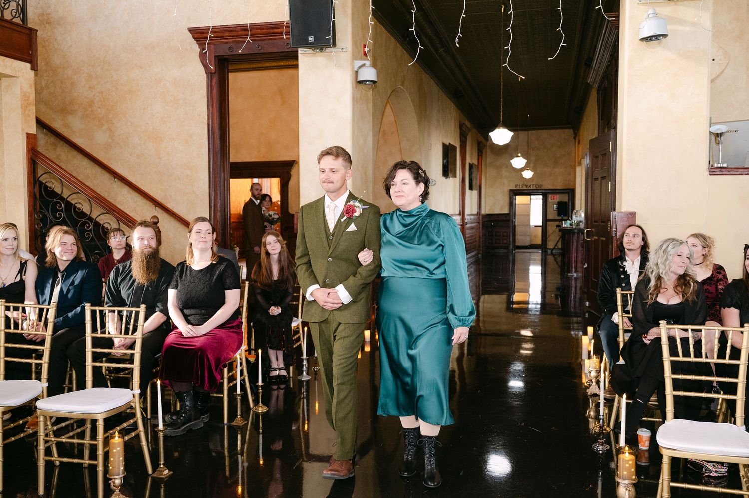 Central MN Wedding Photos | Groom in military uniform walks down wedding aisle with woman in teal dress, guests seated in gold chairs.