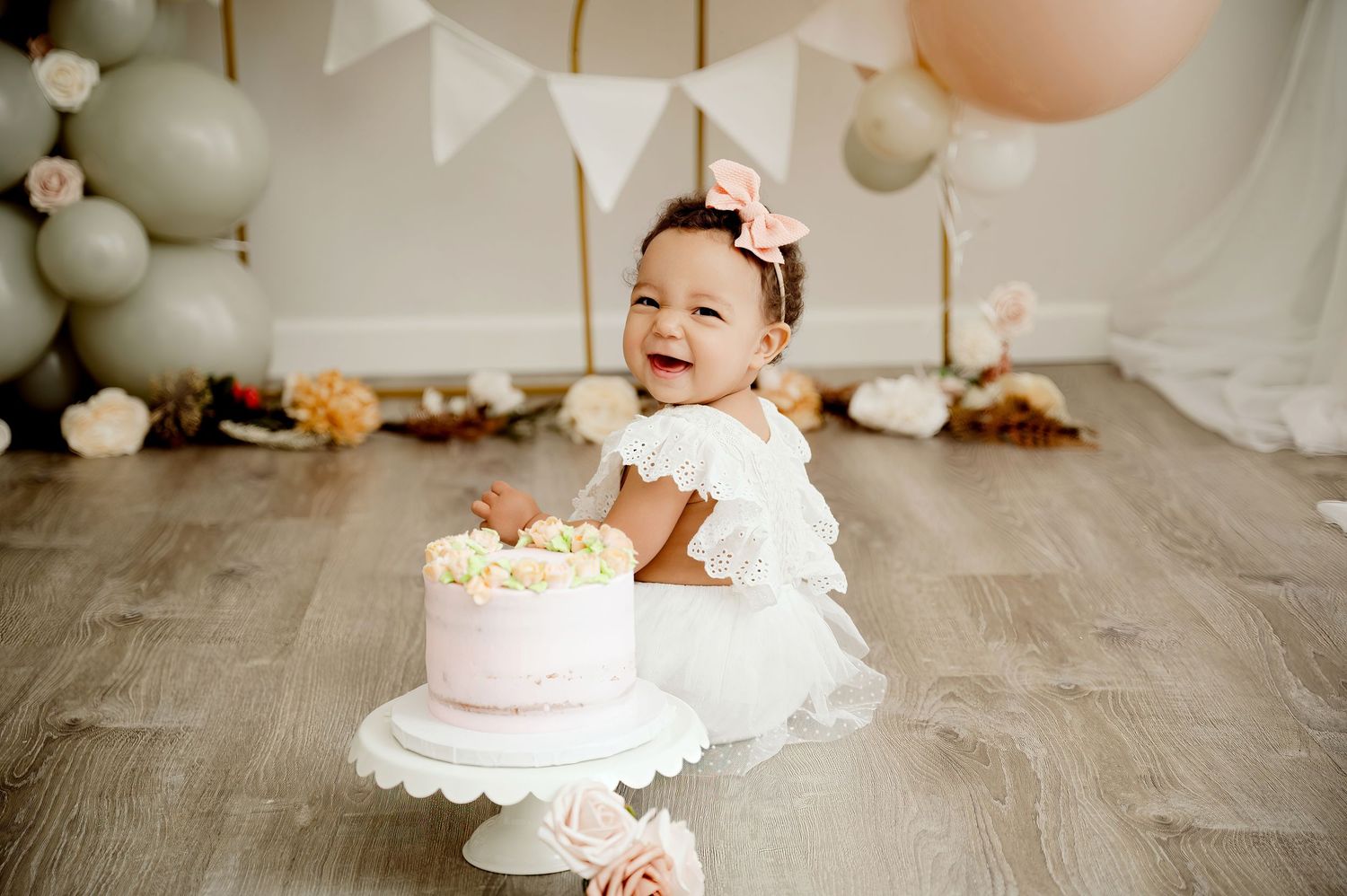Central MN Cake Smash Photos | Baby girl in white dress smiles at her first birthday cake smash with balloon garland backdrop.