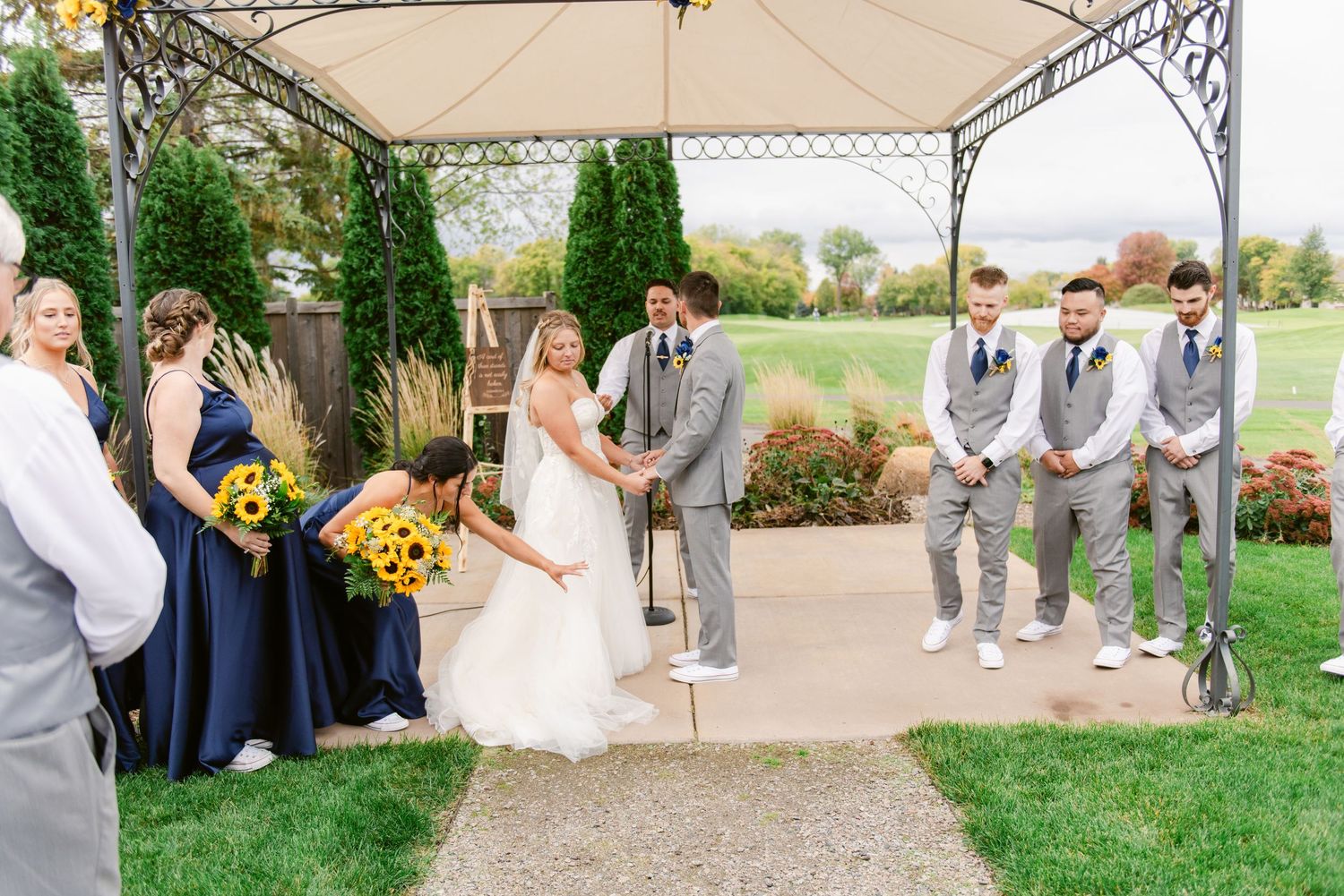 Central MN Wedding Photos - Wedding ceremony under gazebo with bride, groom, groomsmen in gray suits, and bridesmaids in navy dresses.