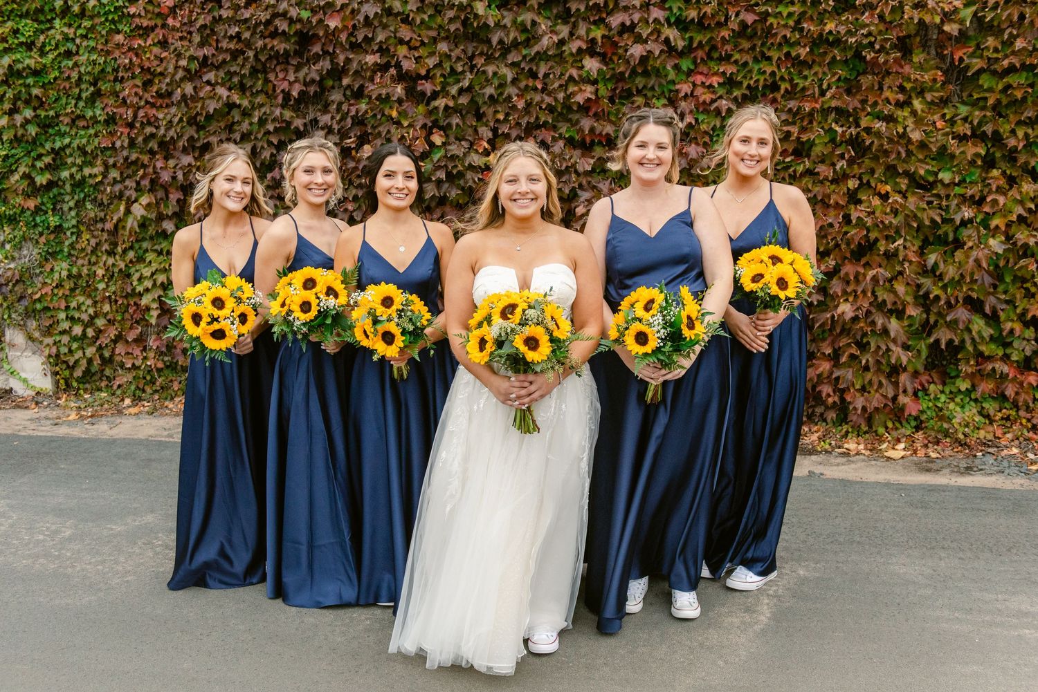Central MN Wedding Photos | Bride with bridesmaids in navy spaghetti-strap gowns holding sunflower bouquets in front of ivy wall.