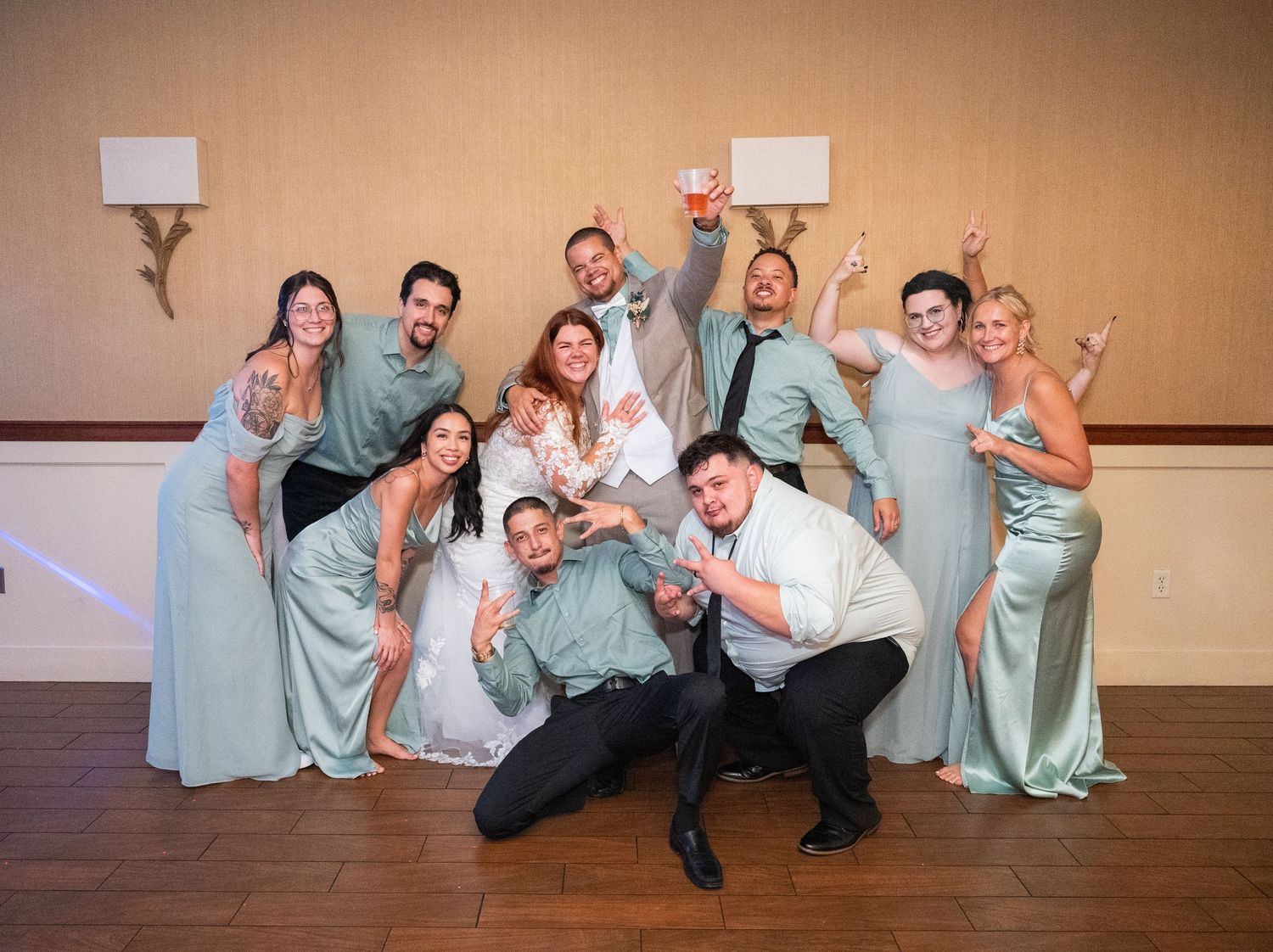 Central MN Wedding Photos | Wedding party in sage green dresses and suits posing playfully together at indoor reception venue.