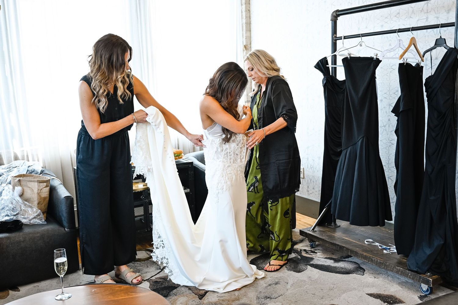 Wedding party members helping with final dress preparations in a bright room.