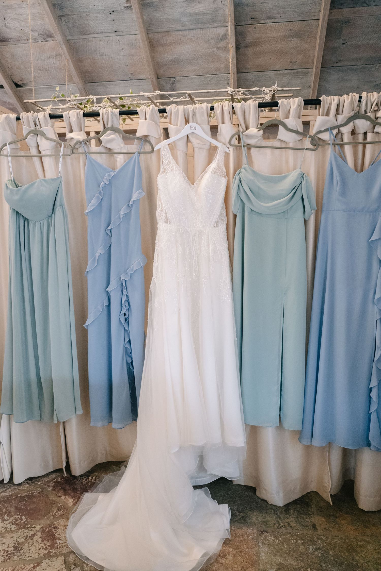 White wedding gown surrounded by light blue bridesmaid dresses hanging on a rack in a rustic venue.