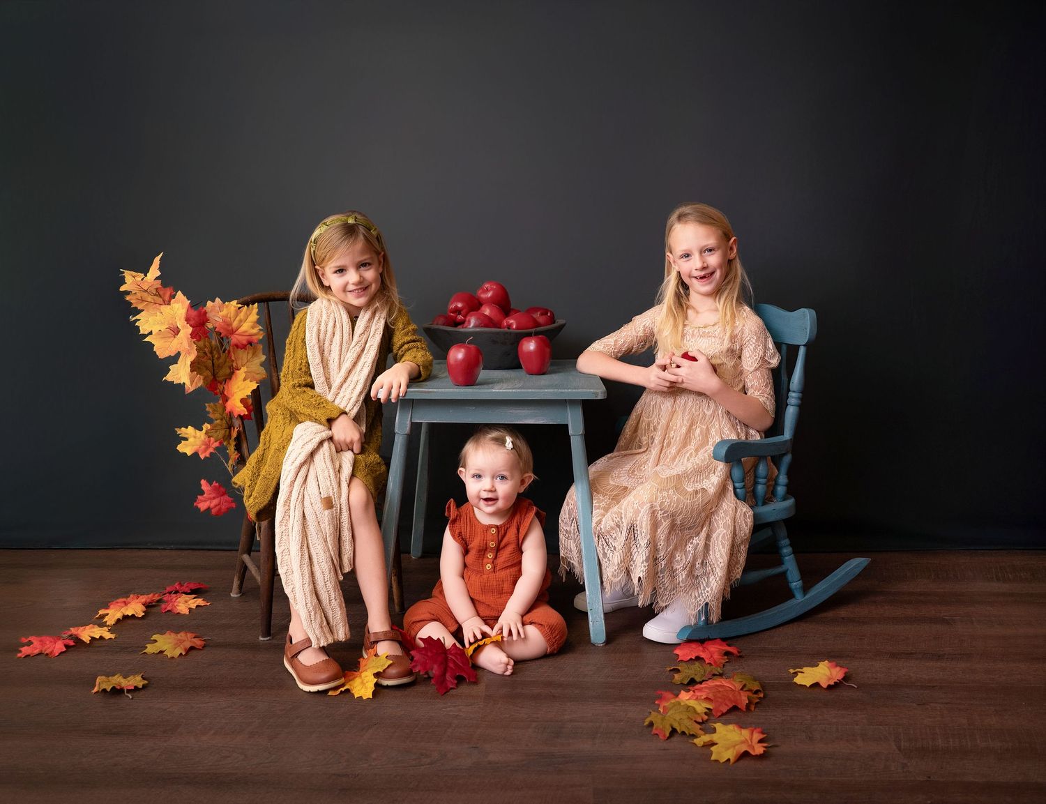A fall-themed studio portrait with autumn leaves, a rocking chair, and apples on a small table against a dark backdrop.