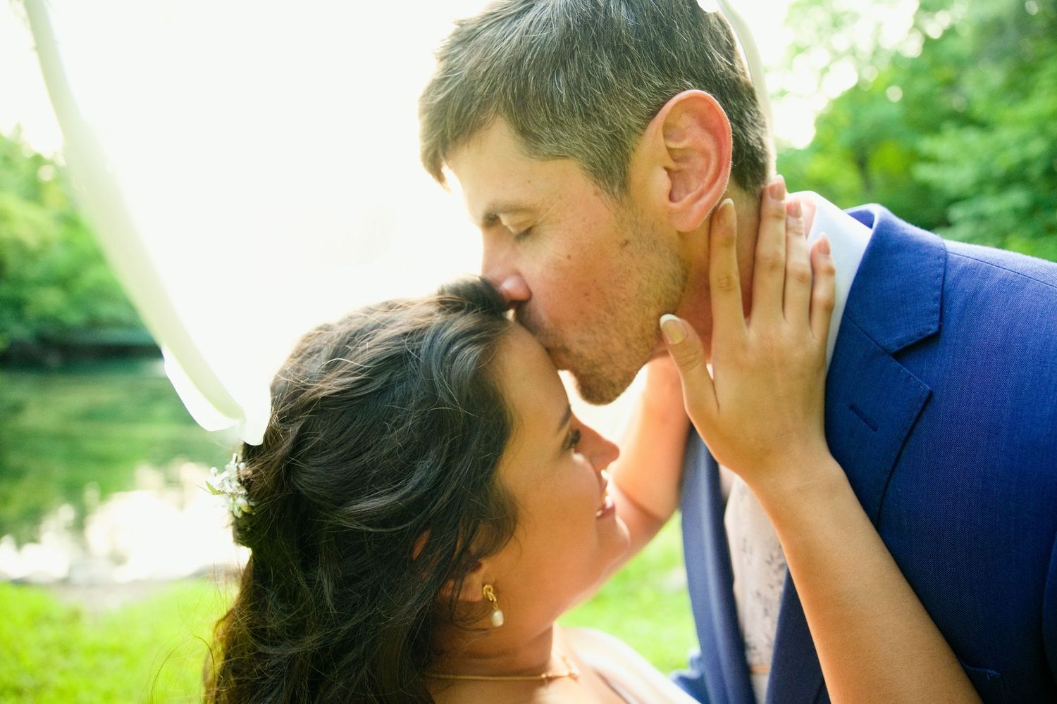 A couple shares a romantic kiss outdoors at their wedding, with lush green trees in the background.