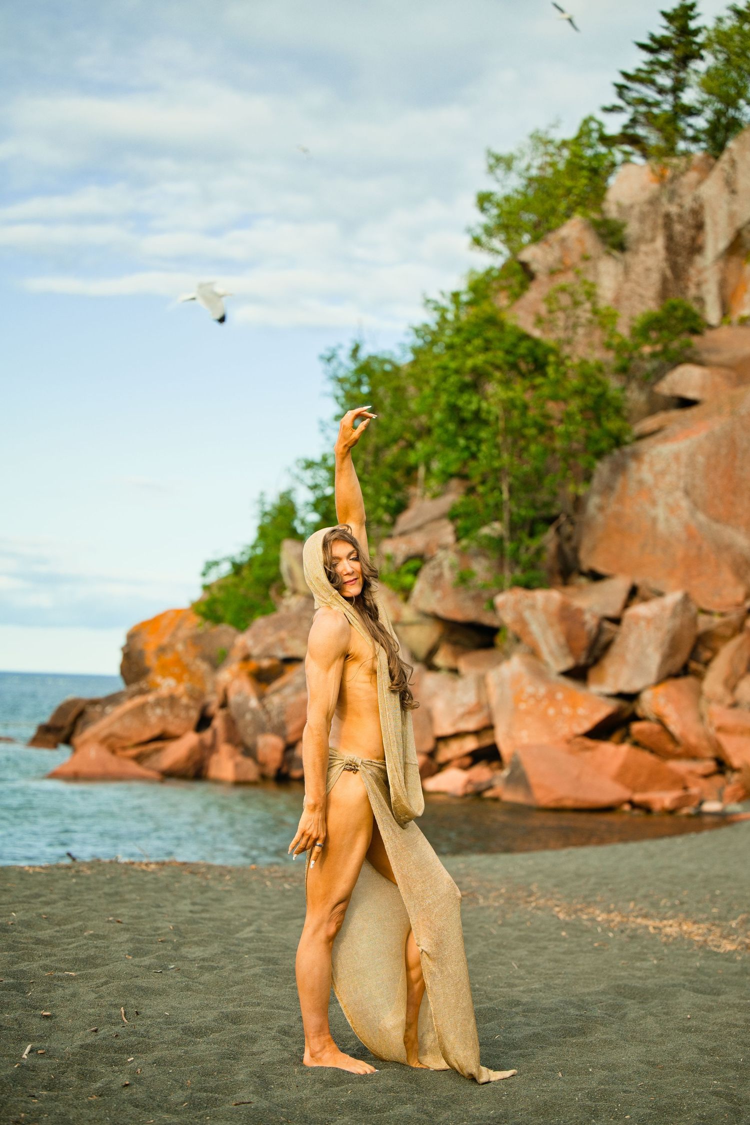 Woman in flowing burlap dress poses on rocky beach shoreline with arm raised, trees and boulders in background.