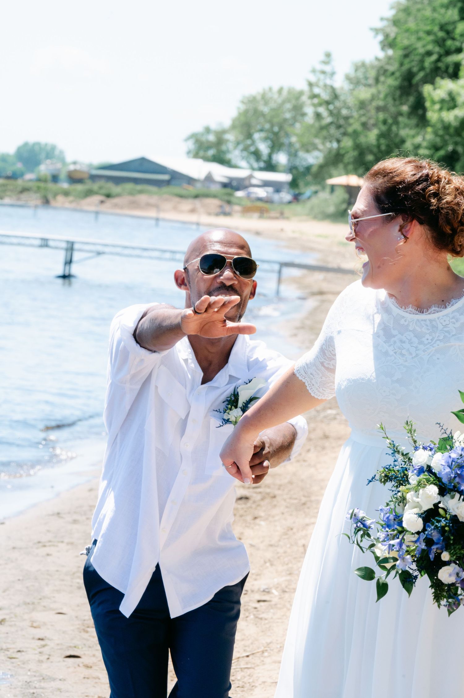 Groom in white shirt and sunglasses laughing with bride in white lace dress holding blue bouquet on sunny beach.