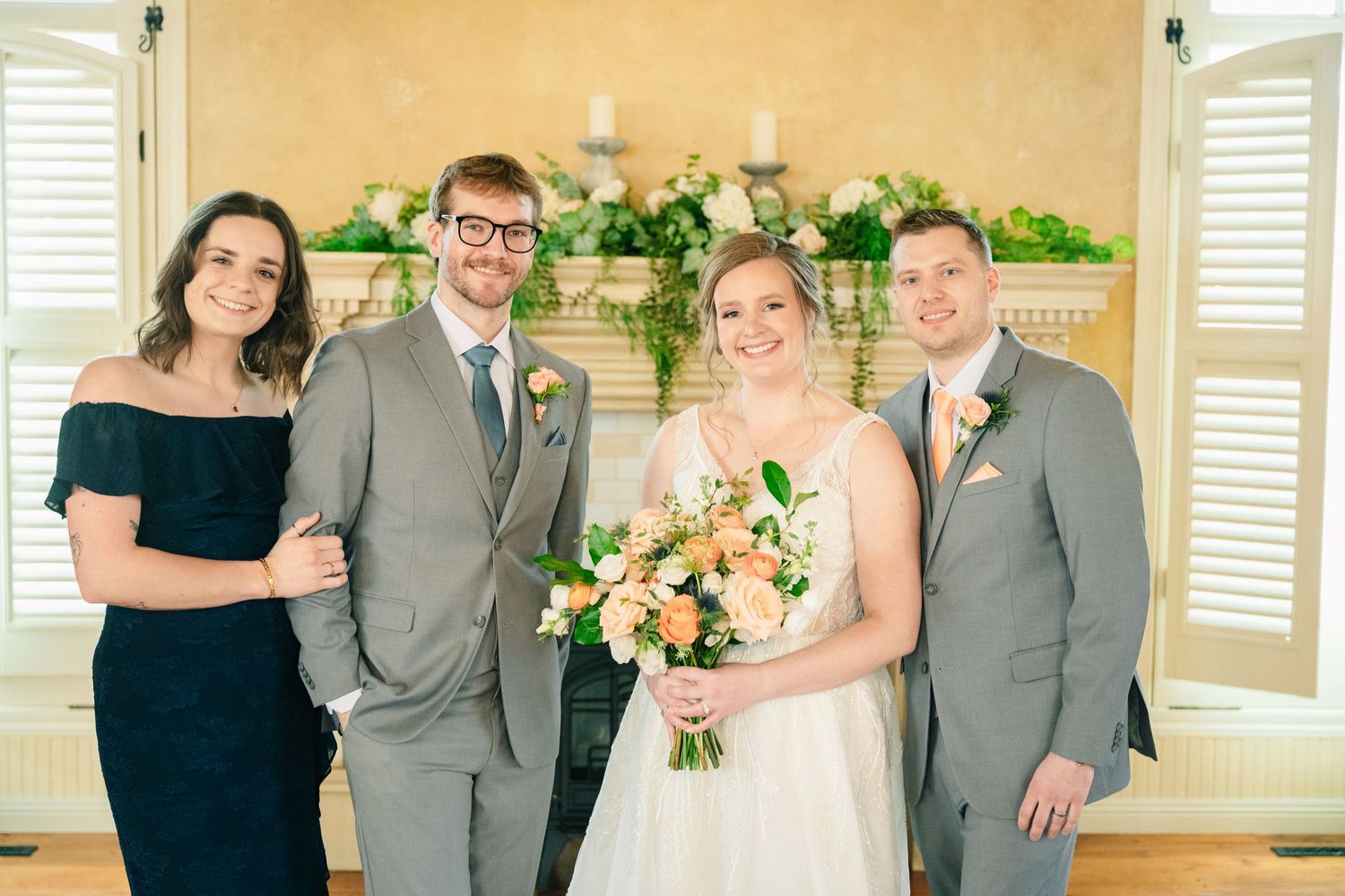 Bride holding peach bouquet poses with groom and two guests in front of decorated fireplace mantel.