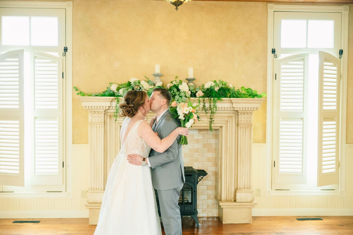 Bride and groom kissing by a floral fireplace mantel in elegant venue.