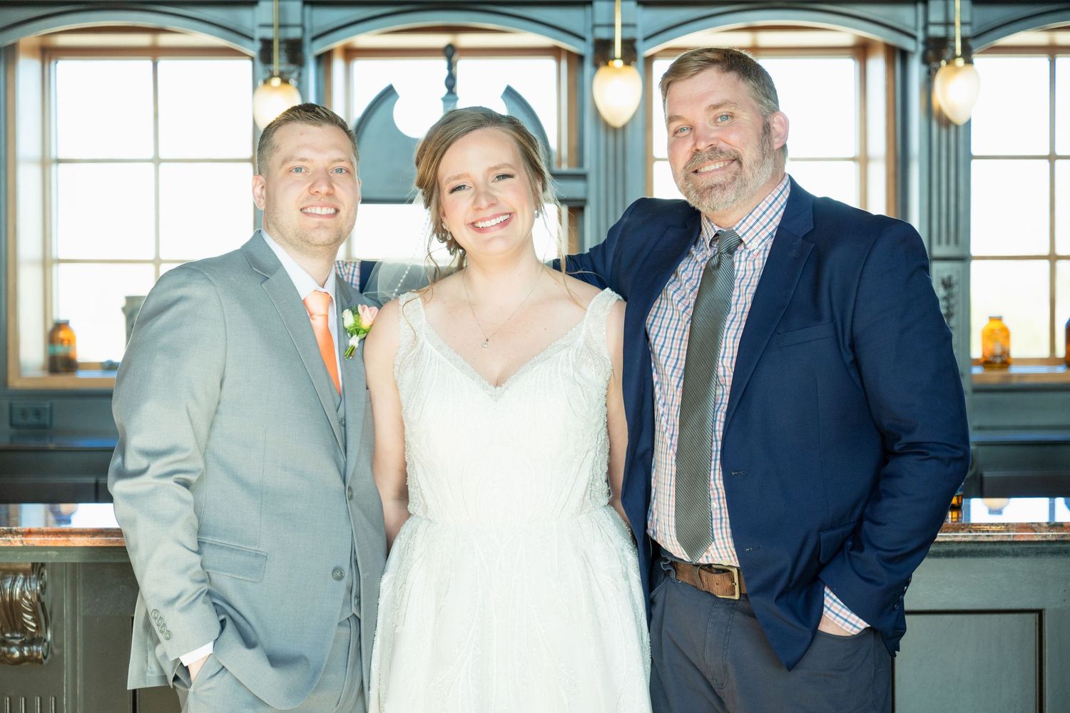 Smiling bride in white gown flanked by two men in suits posing inside rustic bar venue.