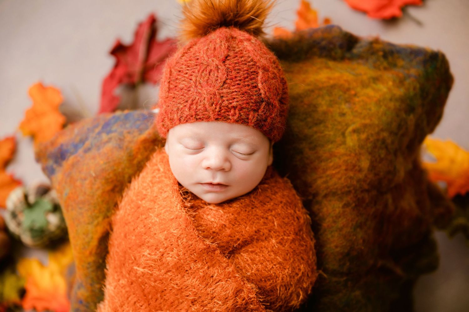 Peacefully sleeping newborn wrapped in cozy orange knit blanket with matching hat among autumn leaves and decor.