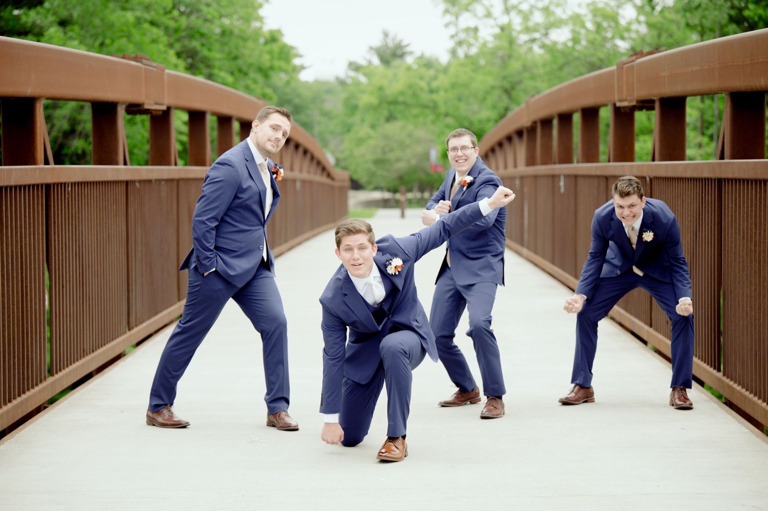 Groomsmen in navy suits pose playfully on wooden bridge during wedding photoshoot.