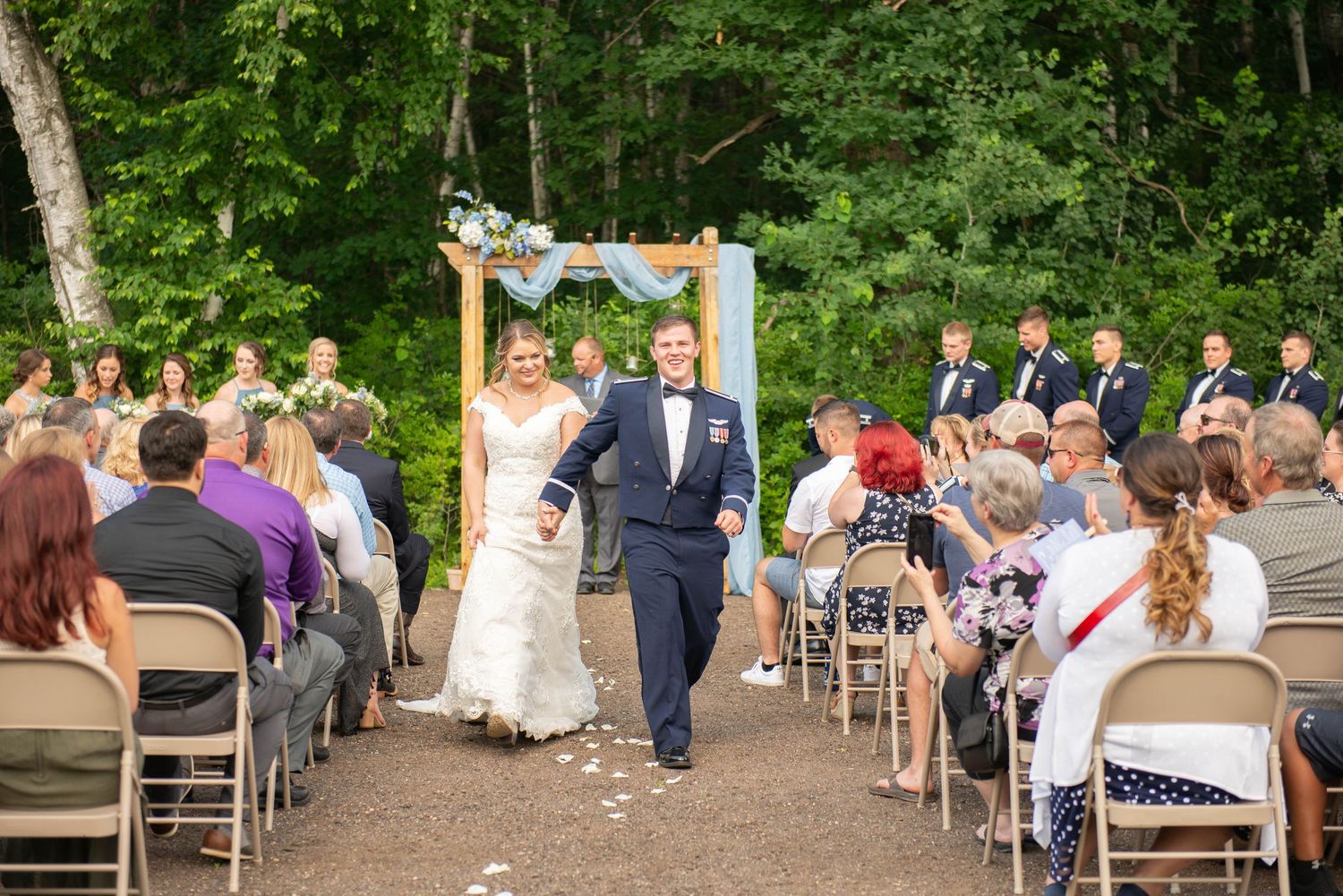 Newlyweds exit their outdoor forest wedding ceremony as guests seated in folding chairs look on. Zimmerman MN Weddings