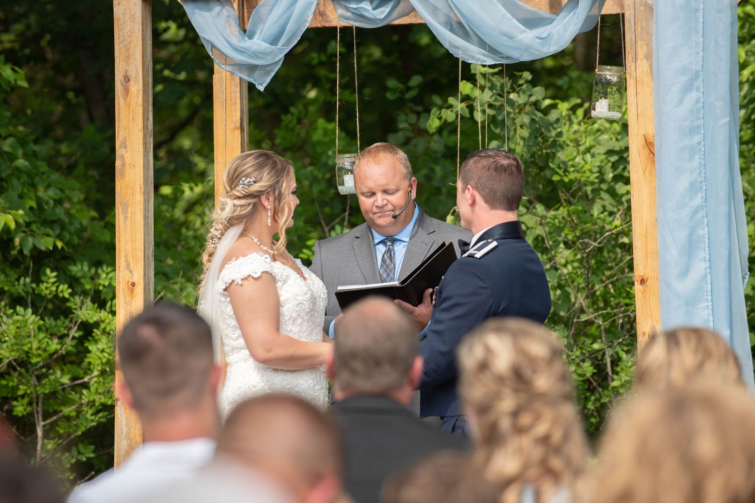 An outdoor wedding ceremony takes place under a wooden arch decorated with light blue fabric and greenery. Zimmerman MN Weddings
