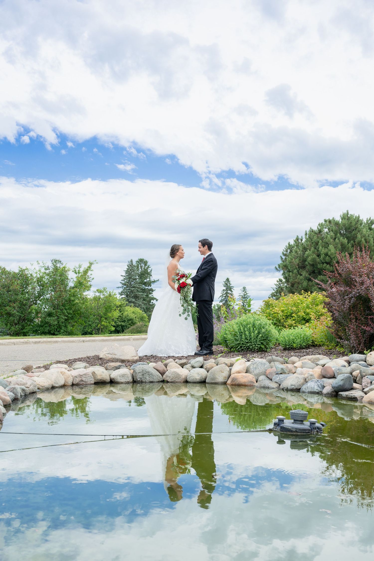 Bride and groom stand by a reflective pond surrounded by lush greenery under a dramatic cloudy sky.