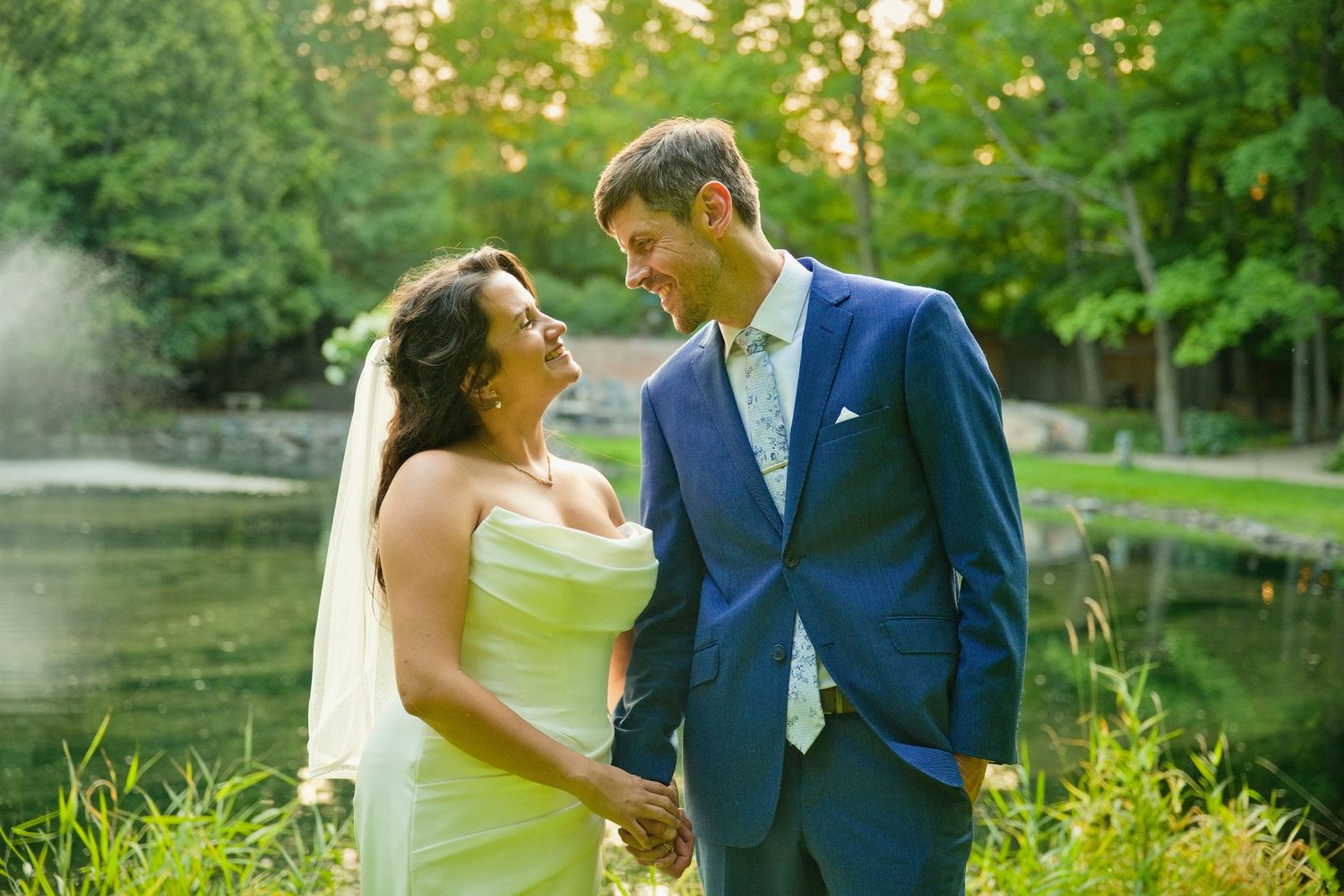 Bride and groom holding hands smiling at each other outdoors near a pond surrounded by lush green trees.