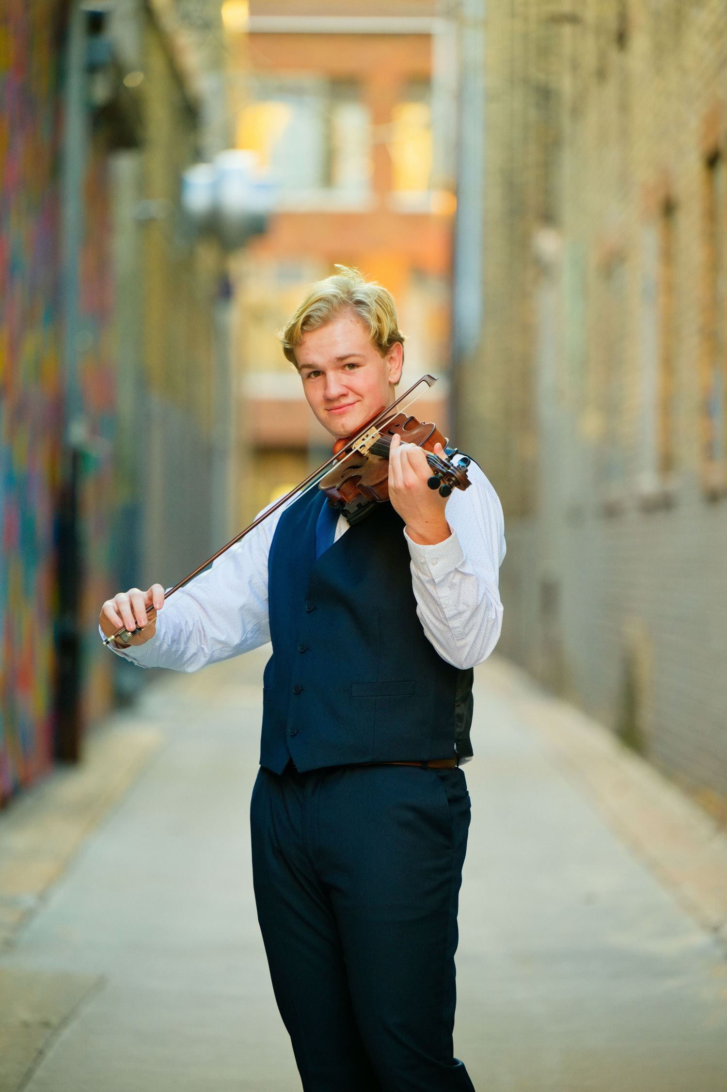 Young male violinist in vest and dress shirt playing violin in urban alley with brick walls.