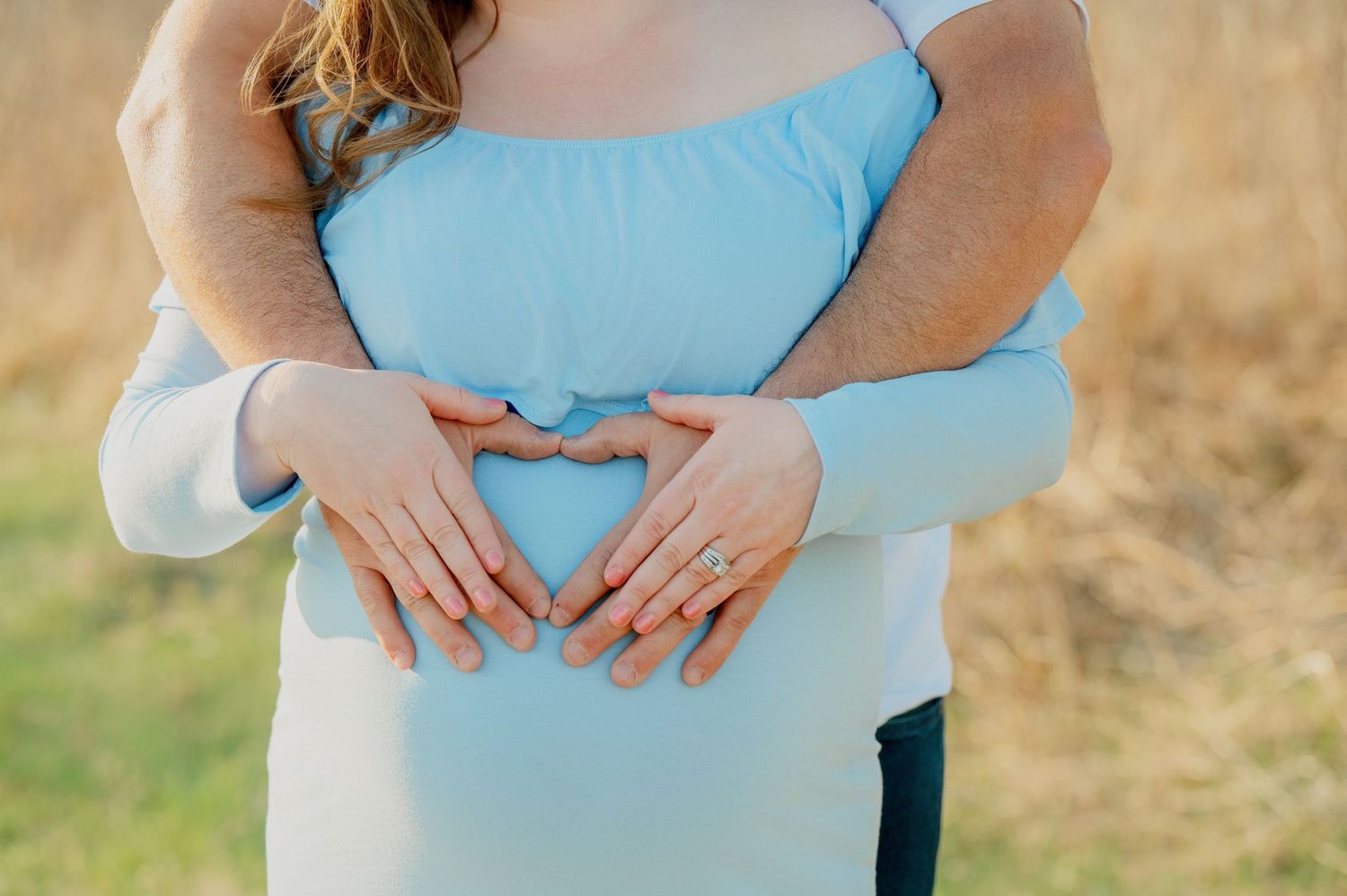 Two sets of hands making a heart shape against a blue maternity dress in a golden field at sunset.