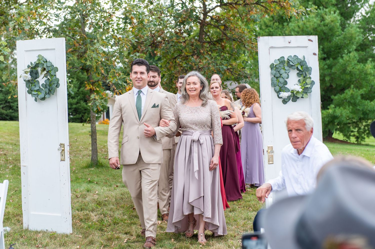 Wedding party walks through an outdoor ceremony space with white doors and floral wreaths on display.