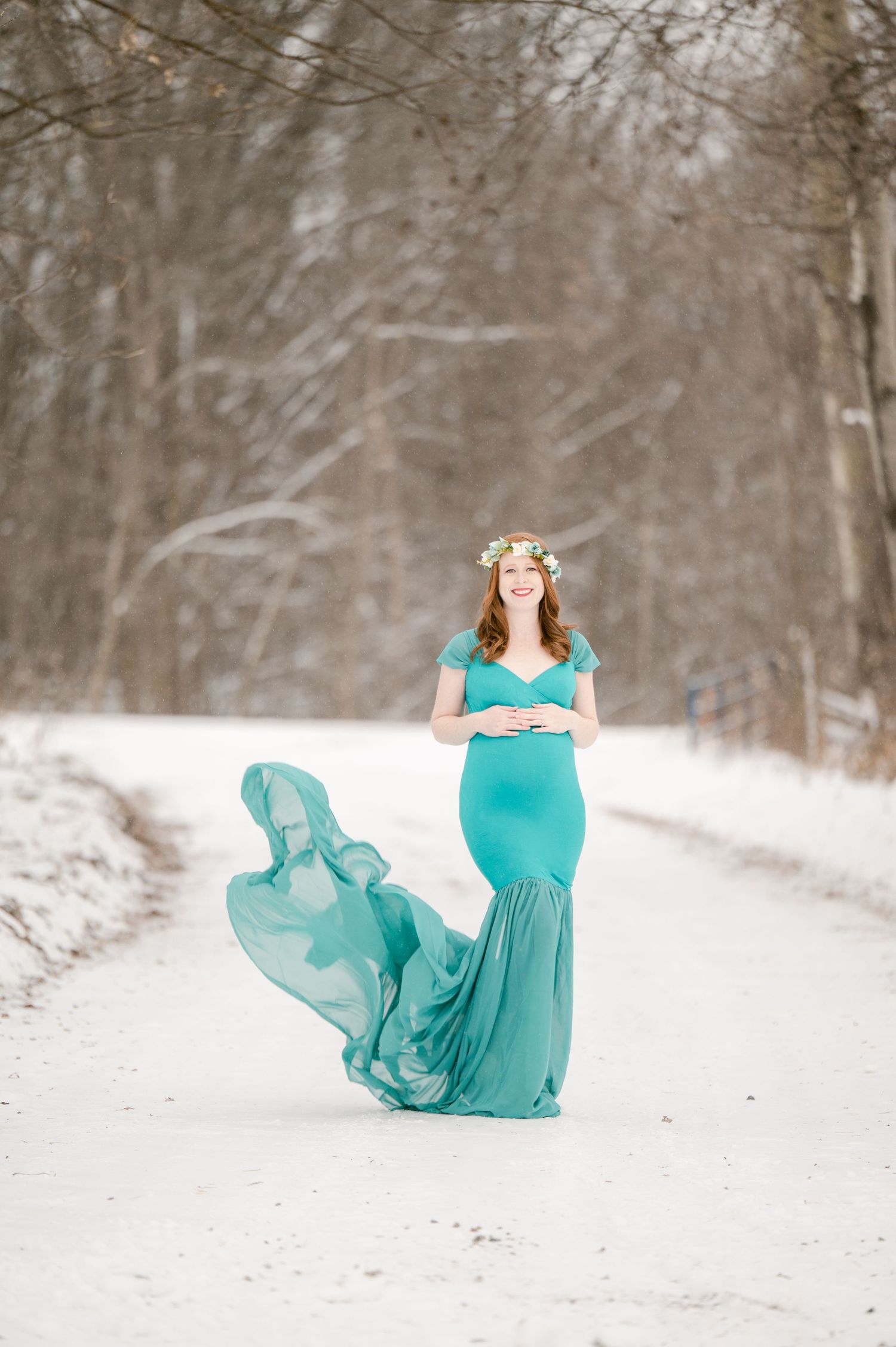 The Clark’s romantic winter maternity photoshoot A woman in a flowing turquoise gown poses gracefully in a snowy winter forest setting.