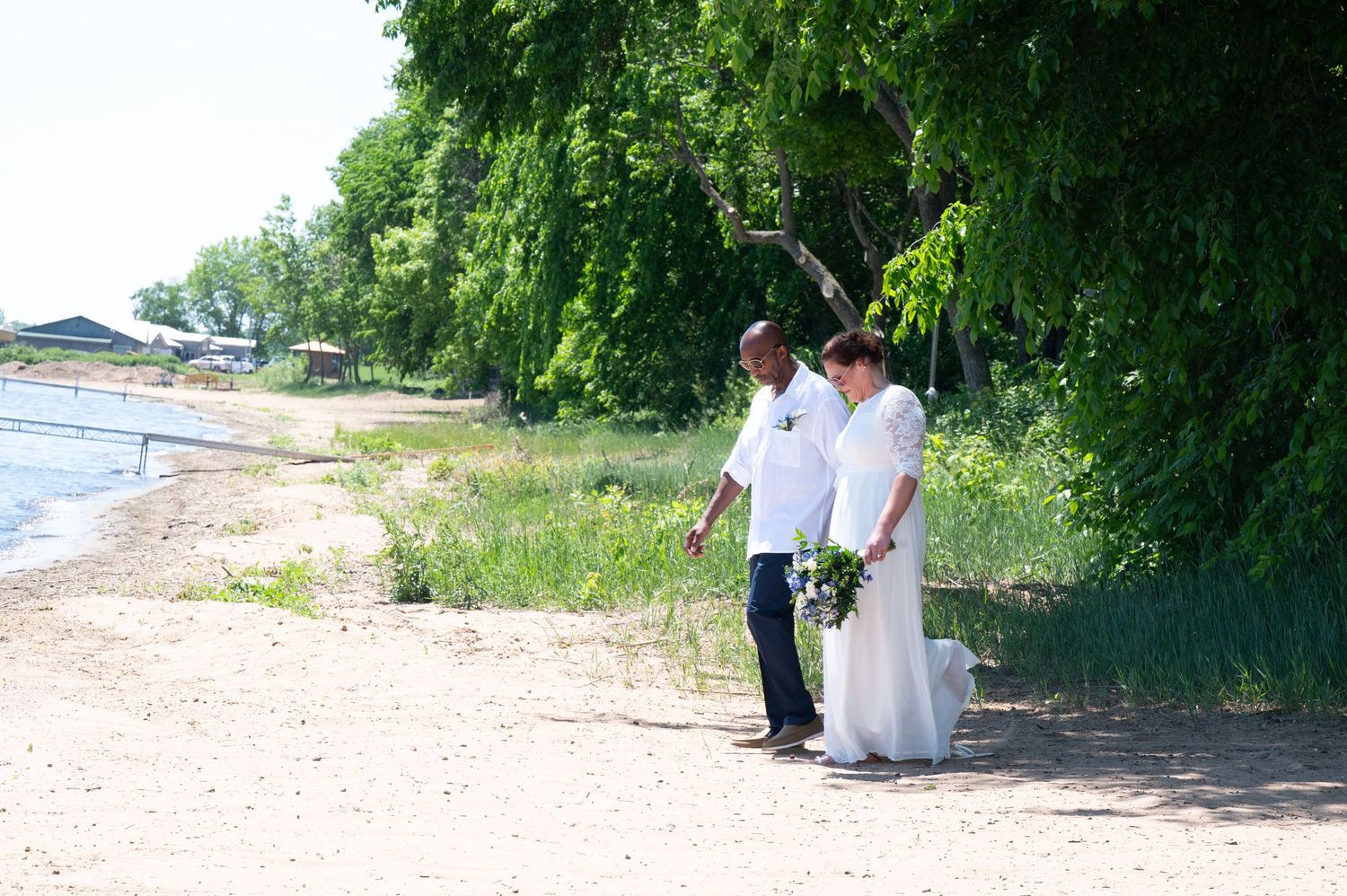 Wedding couple walking along sandy beach shoreline surrounded by lush green trees in summer.