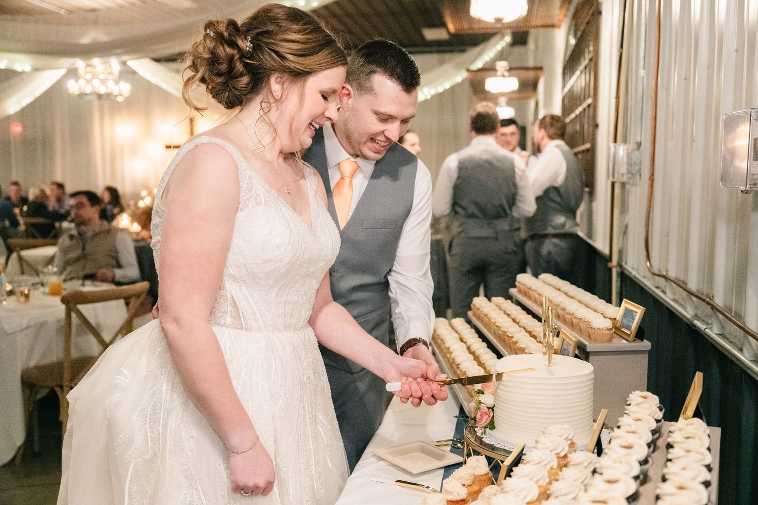Bride and groom laugh joyfully while cutting wedding cake at elegant indoor reception venue.